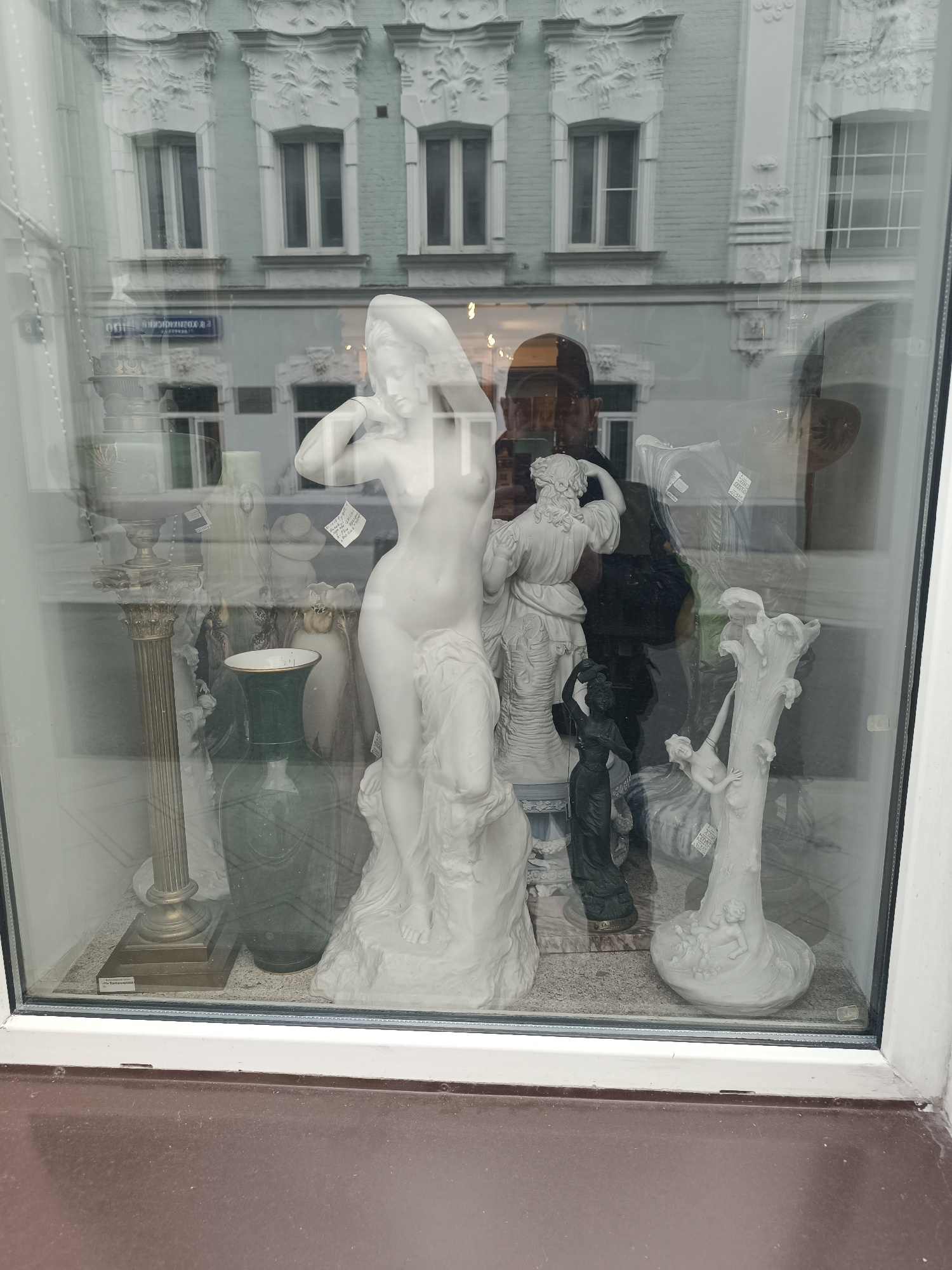 A storefront window displays various classical-style statues and decorative objects. In the foreground, a large white statue of a woman is prominently featured. To its left is a tall, ornate golden pedestal. A dark green vase sits behind the pedestal. Further back, a smaller dark statue of a woman carrying a pot is visible. To the right of the large white statue, a white statue depicting figures entwined with a tree trunk is present. The window reflects the exterior of a building with ornate architectural details and multiple windows. The light is overcast.