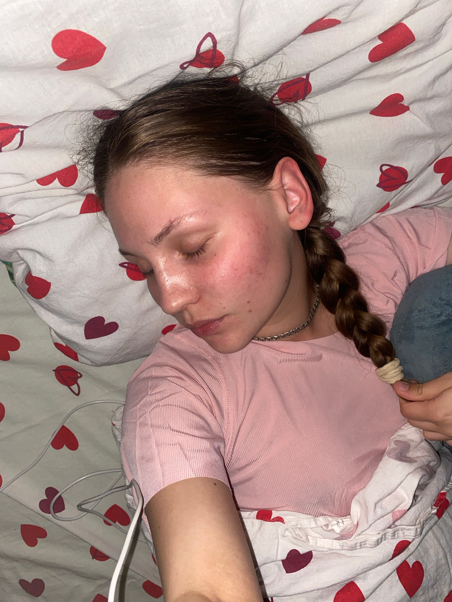 The media file shows a young person sleeping in a bed with a heart-patterned duvet. The person is lying on their side, facing left, with their eyes closed and their face resting on a pillow. They have dark brown hair braided into a single plait, which is secured with a beige elastic band. The person is wearing a pink, long-sleeved top. A greyish-blue plush toy is visible behind their head, partially covered by the duvet. White and grey wires are seen near the person's shoulder, trailing off the right side of the frame. The bedding has a repeating pattern of red and dark pink hearts on a white background. The lighting suggests it might be daytime or a well-lit indoor setting. There is no visible text in the image. The scene conveys a sense of rest and peaceful sleep. The location context provided is Вассята, Russia, which does not offer specific visual cues within the image itself.