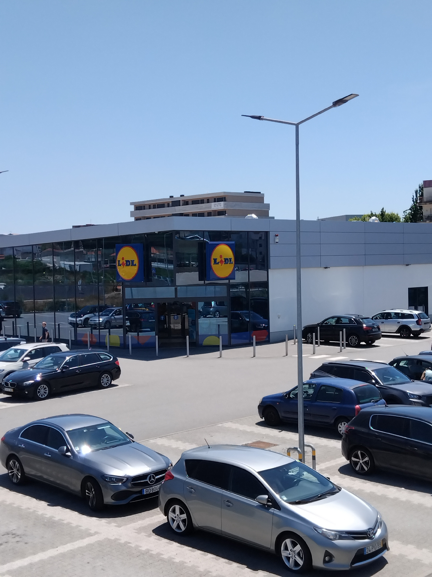The image shows a sunny daytime view of a Lidl supermarket in Vila Nova de Famalicão, Portugal. The supermarket has a modern facade with large glass windows and prominent Lidl logos. In the foreground and surrounding the supermarket are several parked cars of various makes and models. A streetlamp with two lights is visible in the center of the frame, with a clear blue sky above. The ground is paved with what appears to be a parking lot. There are no visible individuals actively engaged in interactions, though a person can be seen walking in the distance near the supermarket entrance. The environment appears to be an urban commercial area.