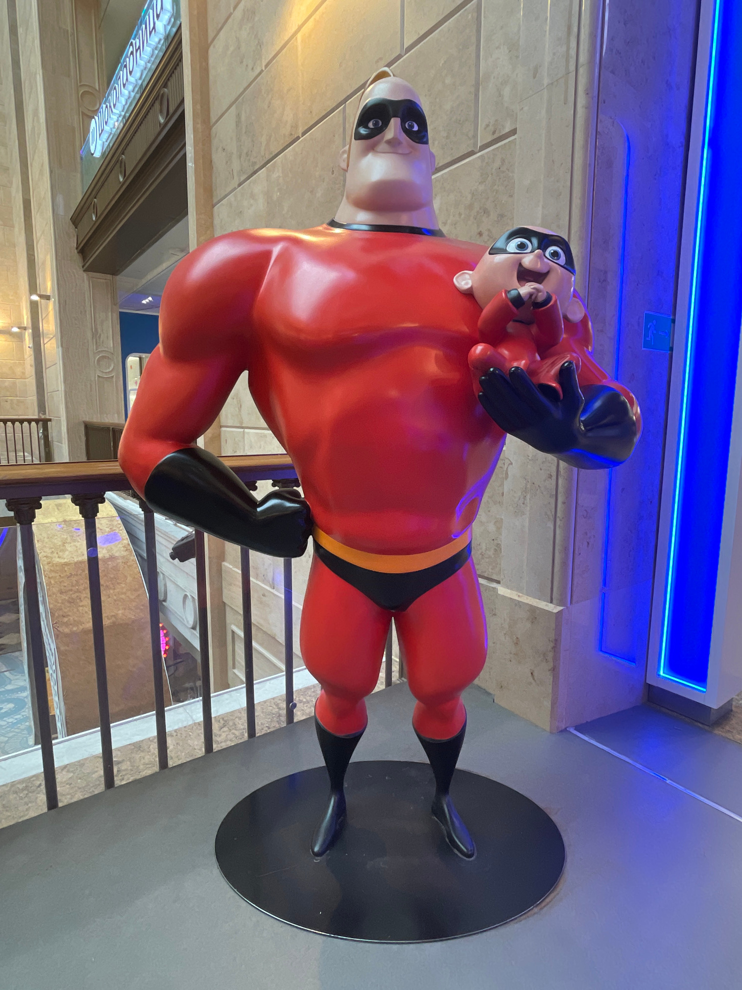 This media file features a statue of Mr. Incredible, a character from Disney's "The Incredibles" franchise. The statue is predominantly red with black accents, including a mask, gloves, and boots. Mr. Incredible is depicted in a dynamic pose, with one arm extended and the other bent at the elbow. He stands on a black circular base.
The statue appears to be located indoors, possibly in a public space like a mall or theme park, indicated by the presence of railings, tiled flooring, and overhead lighting, including a prominent blue neon light strip. In the background, there are glimpses of what might be shops or other attractions.
The statue is a life-sized representation of the character, capturing his muscular physique and heroic stance. The face of Mr. Incredible is detailed, with a determined expression. One of the crops shows a second, smaller Mr. Incredible figure seemingly emerging from the larger one's chest, adding a whimsical or perhaps symbolic element.
There is visible text on a sign in the lower left portion of the image, though it is partially obscured and the text itself is illegible in this orientation. The lighting suggests an indoor environment, and there are no indicators of the time of day or weather. The overall mood conveyed is one of superheroic action and family entertainment.