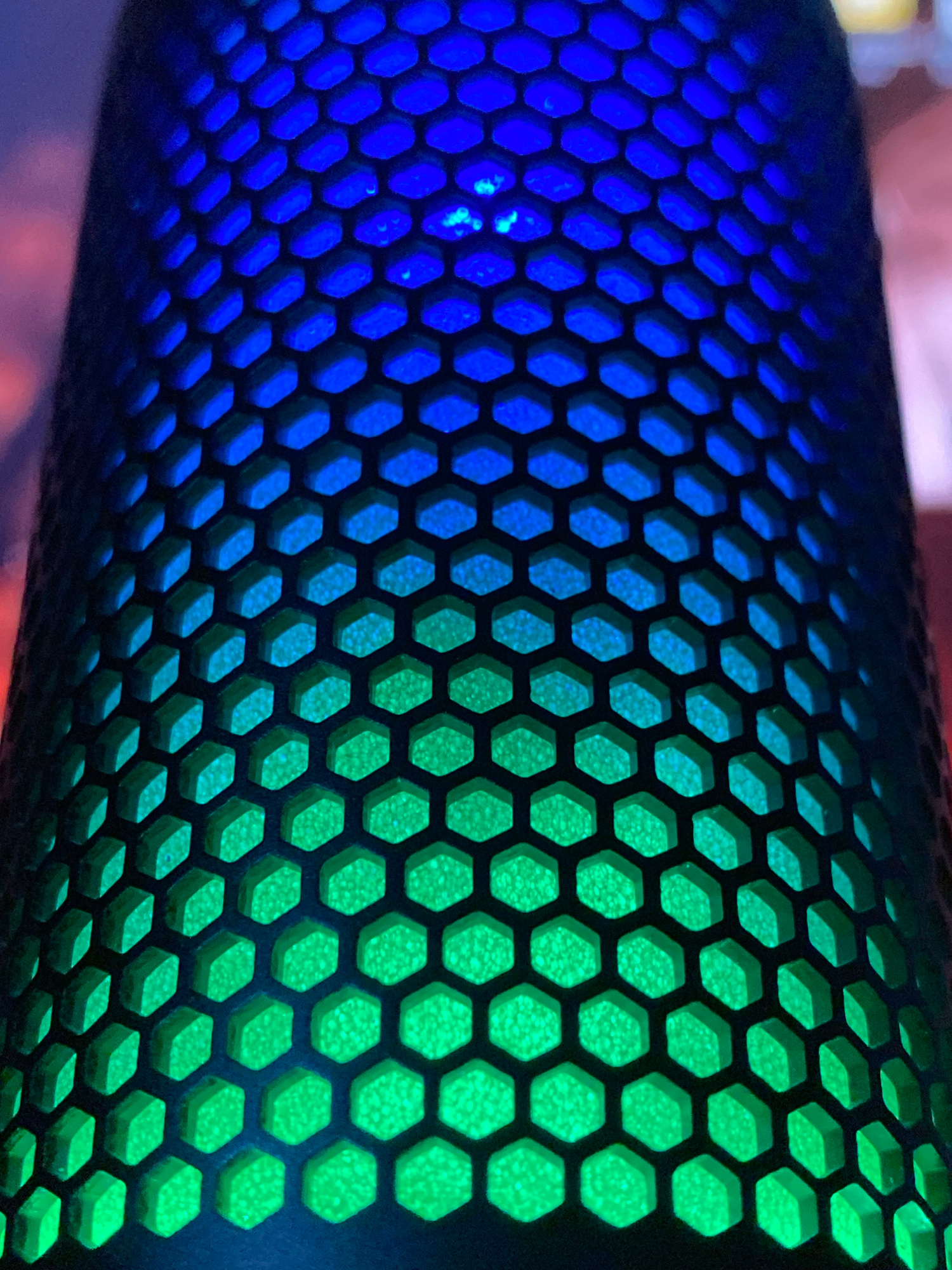 A close-up, vertical shot shows a black, hexagonal mesh with colored lights shining through from behind. The lighting transitions from bright blue at the top of the frame to a vibrant green at the bottom. The hexagonal pattern is distinct and evenly spaced, creating a visually striking gradient effect. The image is cropped tightly on the mesh, with no surrounding environment visible.