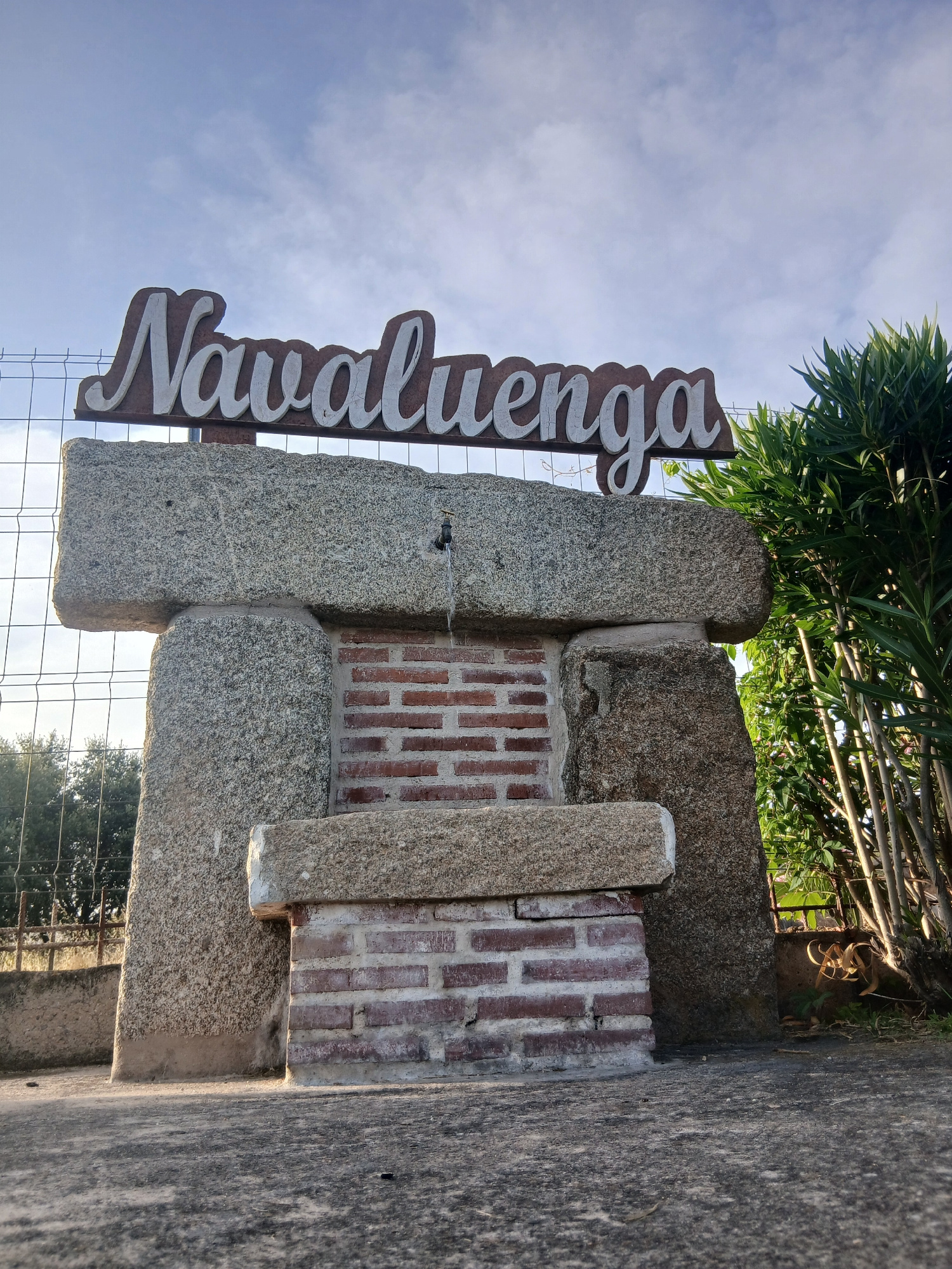 The image shows a stone structure with a sign that reads "Navaluenga." This structure appears to be a public fountain. The sign is wooden with white lettering. The fountain itself is made of rough-hewn granite stones and has a brick backing. A small faucet is visible, and water is flowing from it. The ground in front of the fountain is paved. To the right of the fountain, there are green plants and trees. The background shows a wire fence and more trees, suggesting an outdoor setting. The sky above is blue with some clouds. The image is taken from a low angle, looking up at the fountain and sign. The overall scene suggests a welcoming entrance or landmark in the town of Navaluenga, Spain, known for its natural beauty and historical sites.
