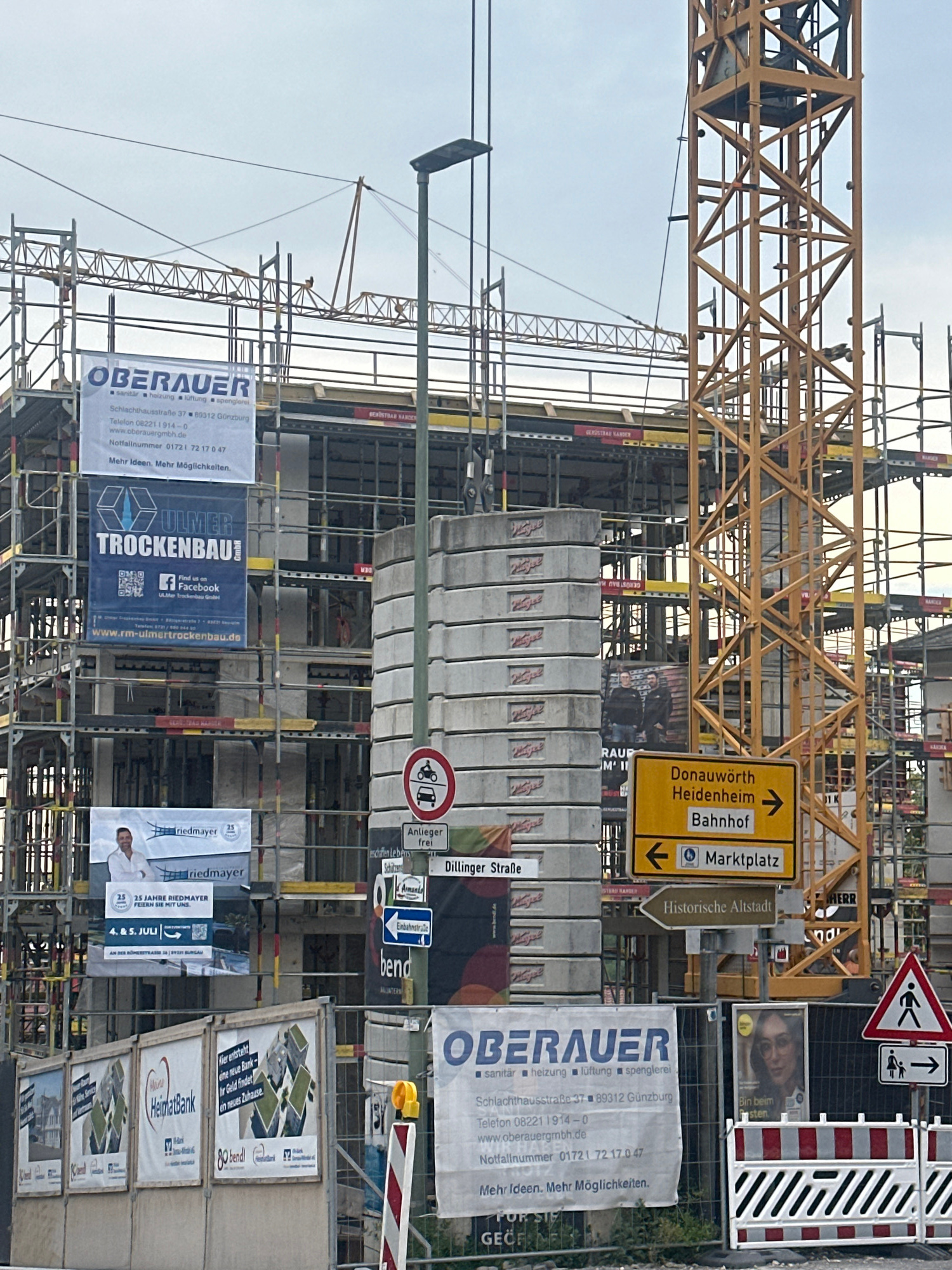 The image captures a construction site with a yellow tower crane prominently featured. The site is a multi-story building undergoing scaffolding and apparent completion. Several banners are visible, advertising construction companies and services. One banner, in blue, promotes "ULMER TROCKENBAU GmbH" with a Facebook link. Another, white banner, displays "OBERAUER" with details about their services (sanitary, heating, ventilation, plumbing) and contact information. A yellow directional sign indicates "Donauwörth Heidenheim Bahnhof" to the right and "Marktplatz" to the left. A smaller brown sign points towards "Historische Altstadt." A traffic sign displaying a motorcycle and car with a red circle and slash indicates that these vehicles are prohibited. Another traffic sign shows a pedestrian with a child and an arrow pointing to the right. In the foreground, there are temporary construction barriers with red and white stripes. The environment appears to be overcast, suggesting a cloudy day. There are no discernible people actively engaged in construction activities within the frame.