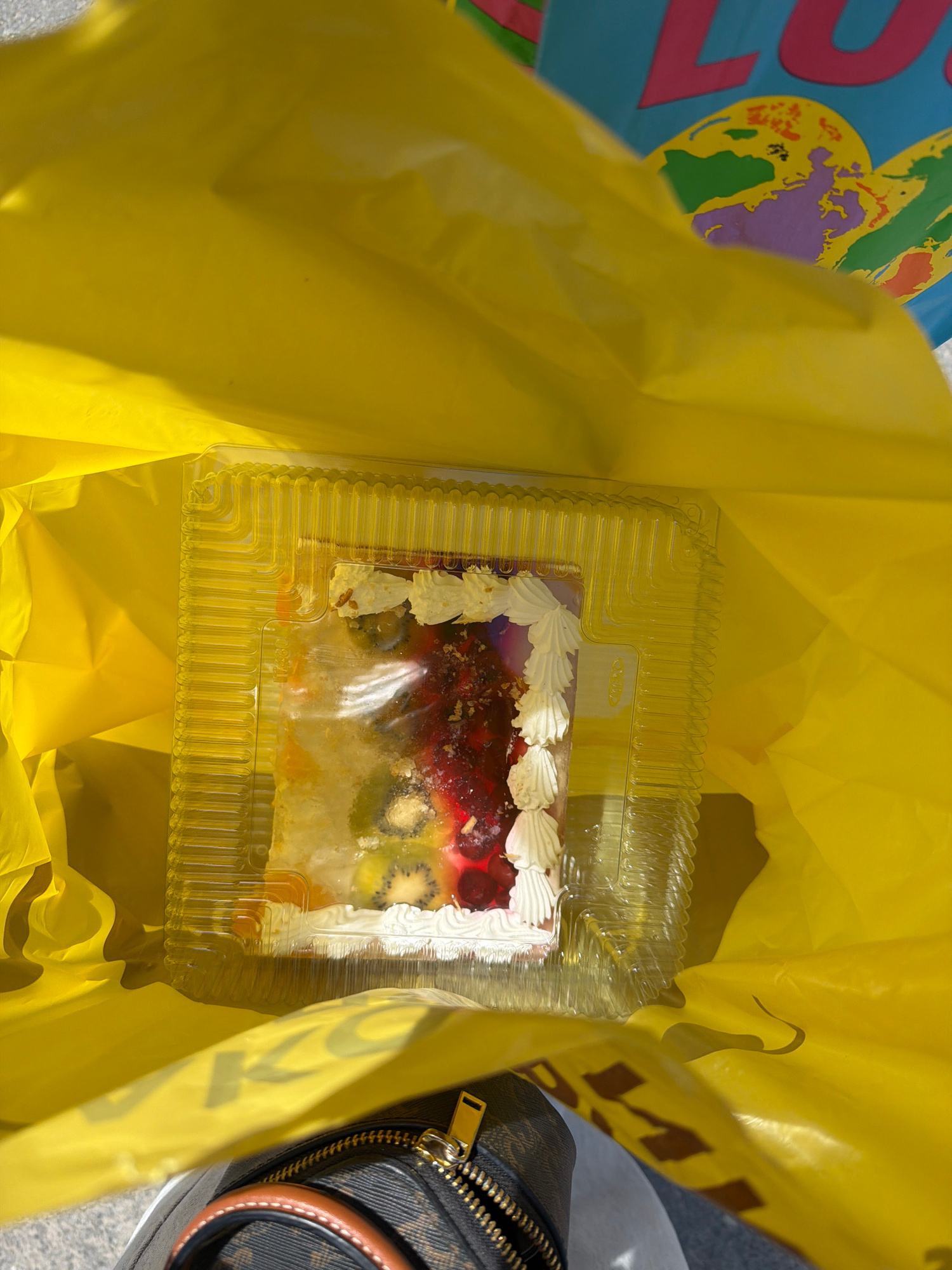 A slice of fruitcake is pictured inside a clear plastic container, which is then placed within a bright yellow shopping bag. The cake appears to have a white frosting, red berries, and slices of kiwi visible. Part of the bag is open, revealing a portion of a blue bag with graphics of the Earth and the word "LU" in large pink letters. A brown and black patterned handbag with a gold zipper is visible at the bottom of the frame. The scene appears to be outdoors, possibly on a street or sidewalk, given the light conditions and a hint of pavement in the upper left corner. The location is identified as Dnipro, Ukraine.