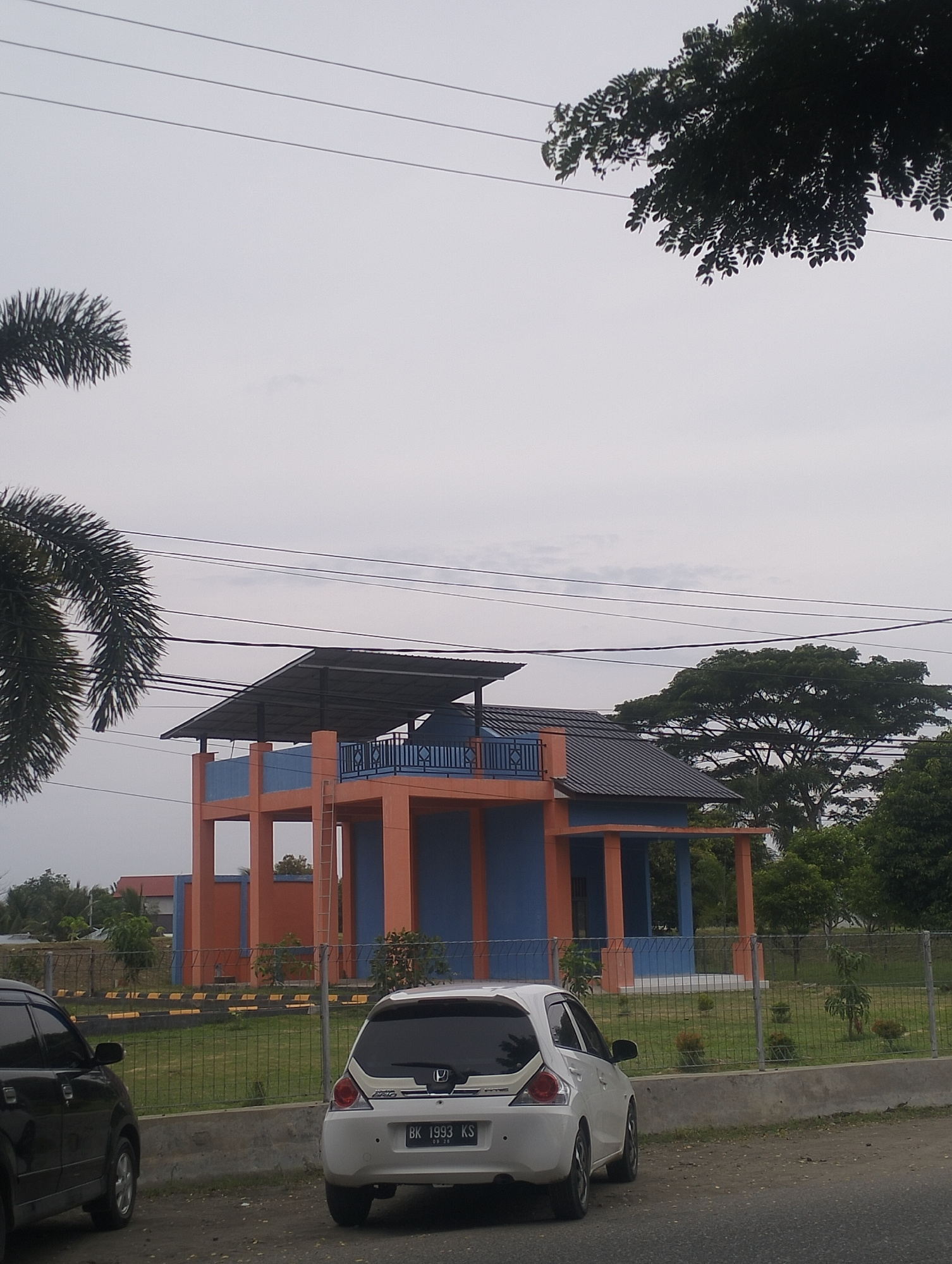 A low-angle, full shot captures a modern, two-story building with orange pillars and blue walls, topped by a slanted metal roof. The structure is fenced, with a grassy yard and scattered trees. In the foreground, a white hatchback car with license plate "BK 1993 KS" is parked. To the left, the rear of a black car is visible. Overhead, power lines crisscross against a hazy sky. The scene appears to be in Lhokseumawe, Indonesia.