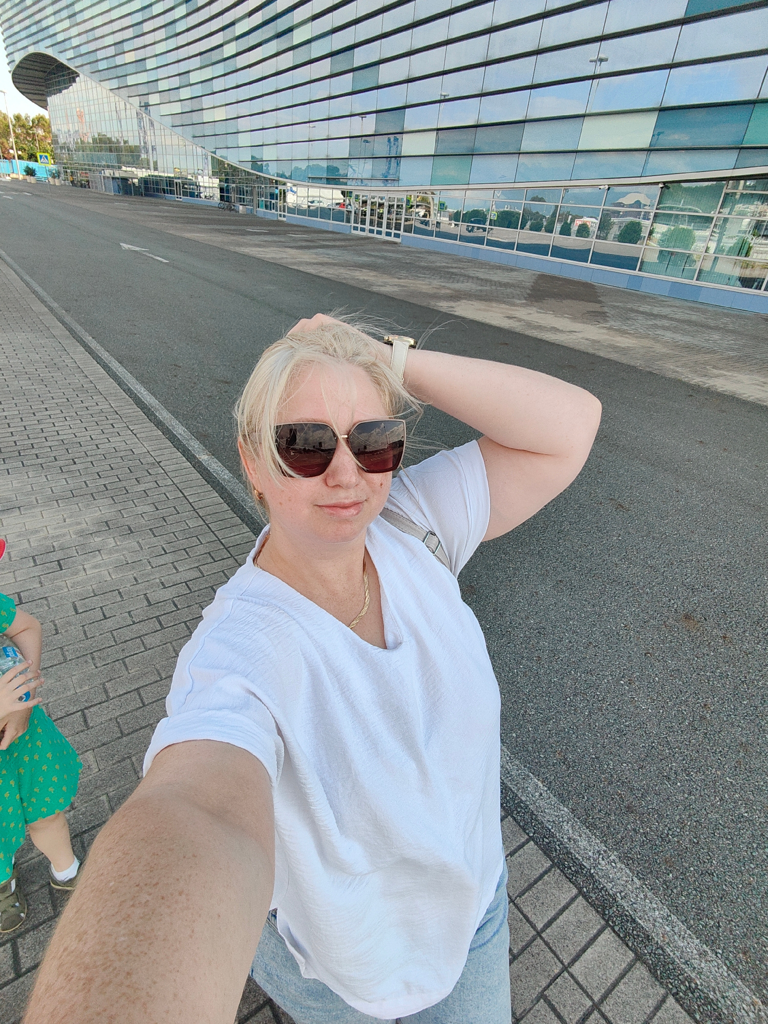 The image captures a woman taking a selfie outside a modern building with a distinctive curved facade, located in Sochi, Russia, likely near an Olympic venue due to the architecture. She has blonde hair, wears large sunglasses and a white V-neck t-shirt. Her right arm is raised, with her hand touching her head. A small child in a green dress is partially visible to her left, holding a bottle. The environment includes a paved sidewalk, a road with lane markings, and the glass-paneled building reflecting the sky and surrounding trees. The lighting suggests it is daytime.