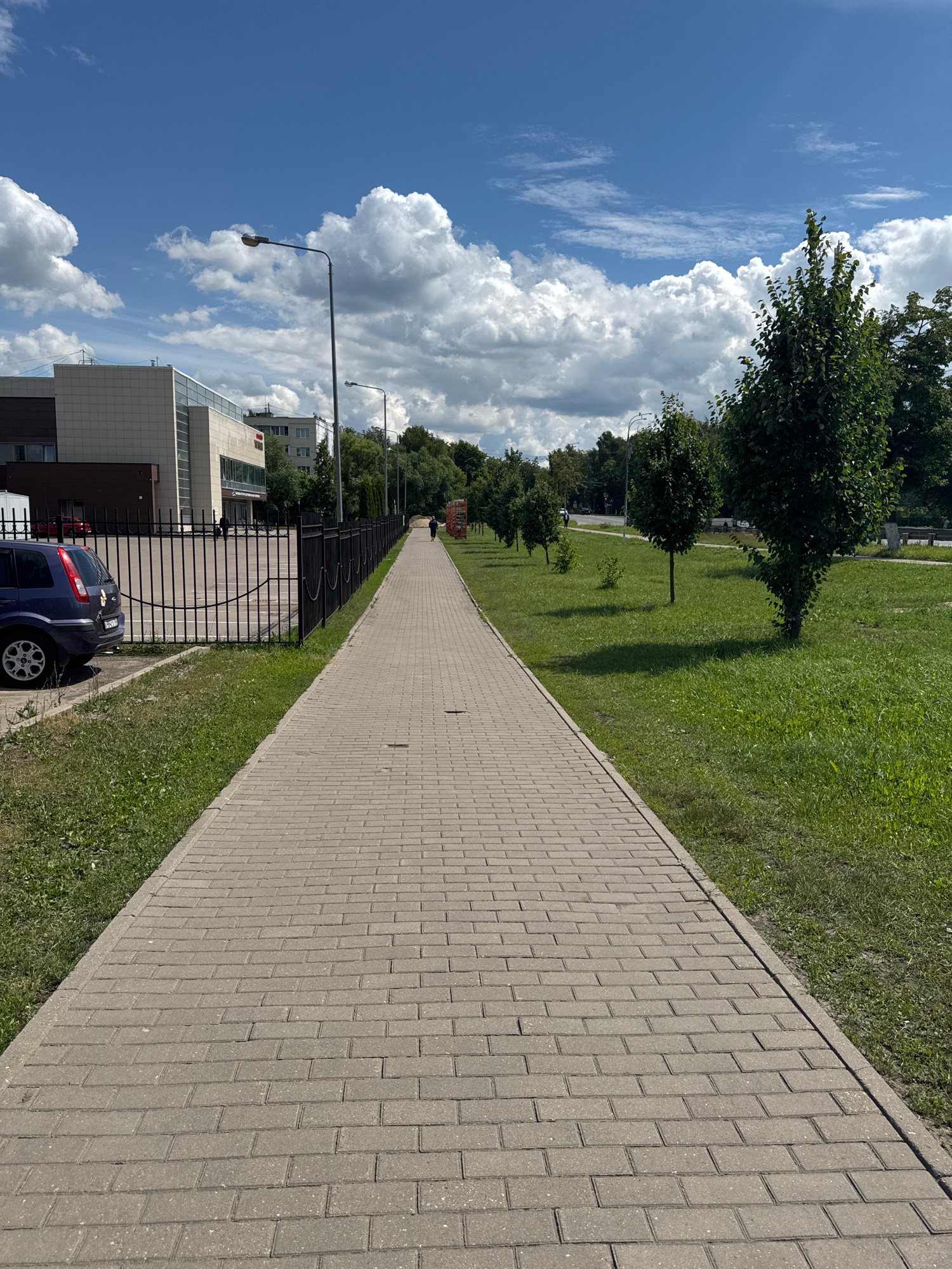 A paved walkway made of light-colored rectangular bricks stretches into the distance, bordered by a grassy area on the right and a black metal fence on the left. A blue car is parked to the left of the walkway, near a modern-looking building with a light-colored facade and dark trim. Beyond the fence, there are several streetlights and trees. The sky is blue with scattered white cumulus clouds. A lone figure is visible walking away from the camera down the walkway, and another person is walking further in the distance. The scene appears to be a public space, possibly a park or a commercial area, on a bright, sunny day. This image was taken in the city of Bronnitsy, Russia.