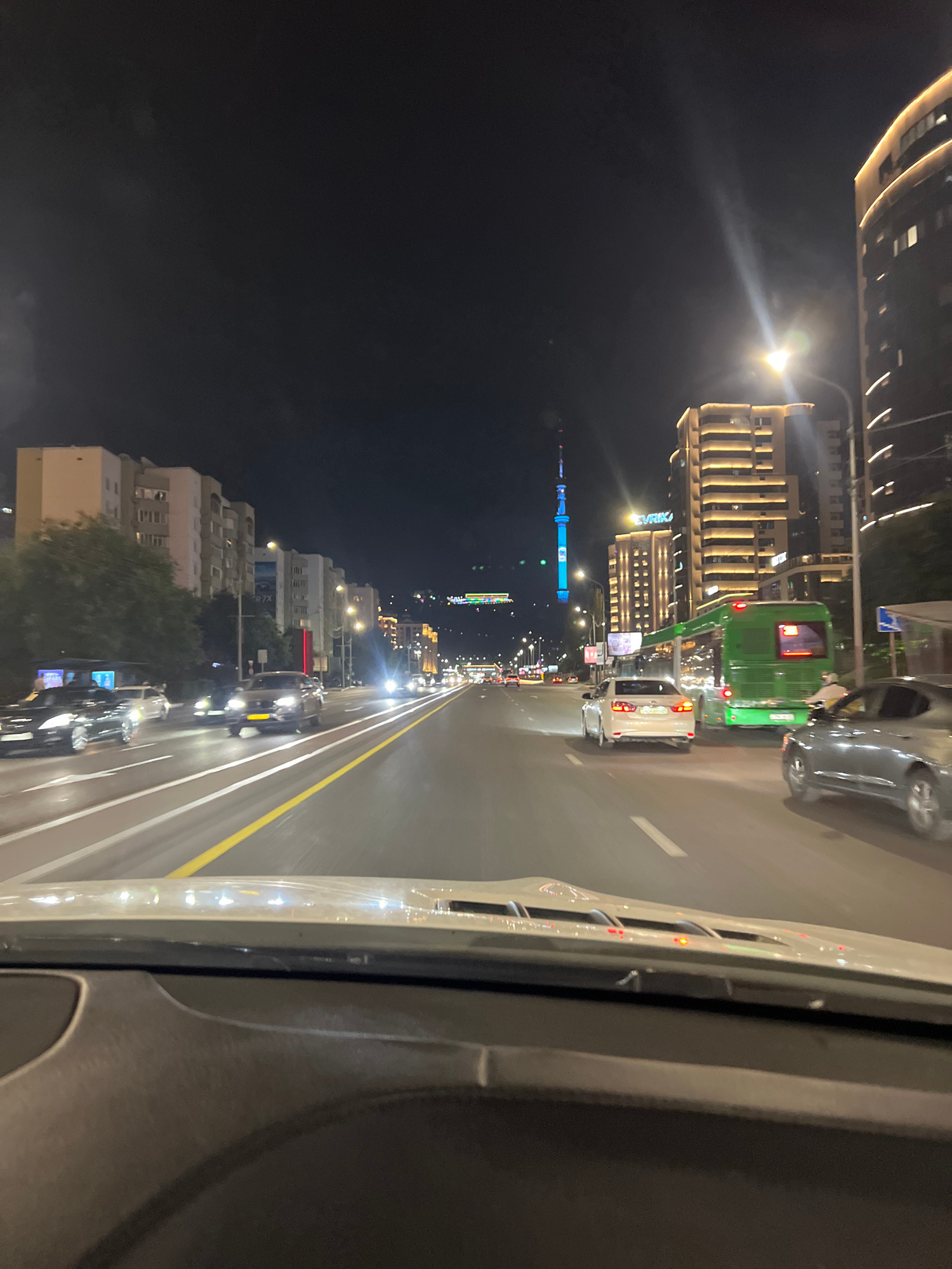 This is a night time, eye-level, forward-facing shot from inside a vehicle driving on a multi-lane road in Almaty, Kazakhstan. The road is lit by streetlights and the headlights of numerous vehicles, including a green bus in the right lane and several passenger cars. Buildings with illuminated windows line the right side of the road, featuring modern architecture and glowing signs. On the left side, there are apartment buildings and trees. In the distance, a tall, illuminated tower, likely the Almaty TV Tower, stands against the dark sky. The vehicle's dashboard is partially visible in the foreground.