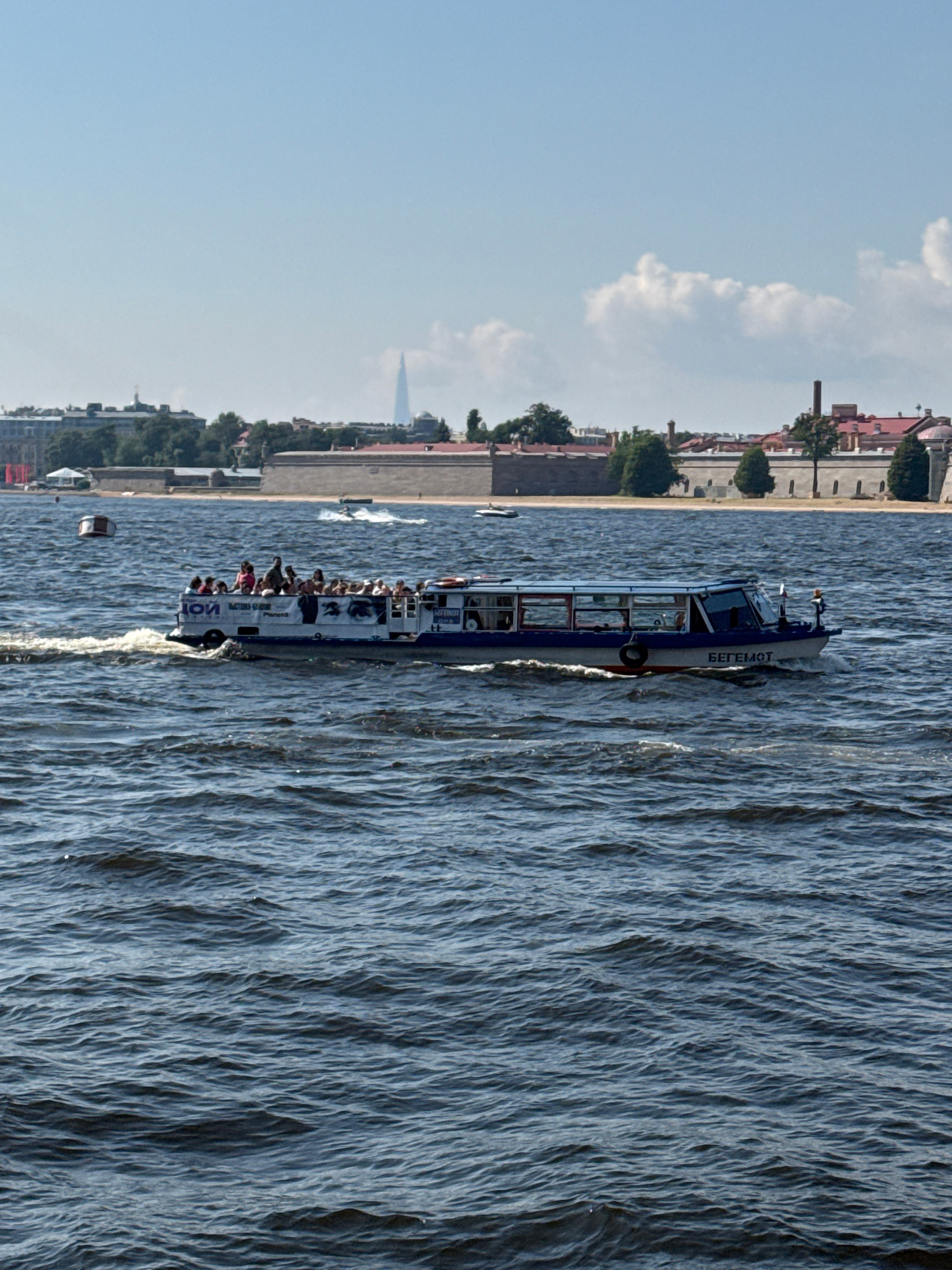 The image depicts a passenger boat named "BEGEMOT" cruising on a body of water, likely the Neva River in Saint Petersburg, Russia. The boat is filled with passengers, many standing and observing the surroundings. The boat has a blue and white hull with windows along its side. In the background, the cityscape of Saint Petersburg is visible, characterized by historical buildings with red tiled roofs and a distinctive tall, slender spire in the distance, possibly the Lakhta Center. The water is choppy, indicating a breezy day. The sky is clear with a few scattered clouds.