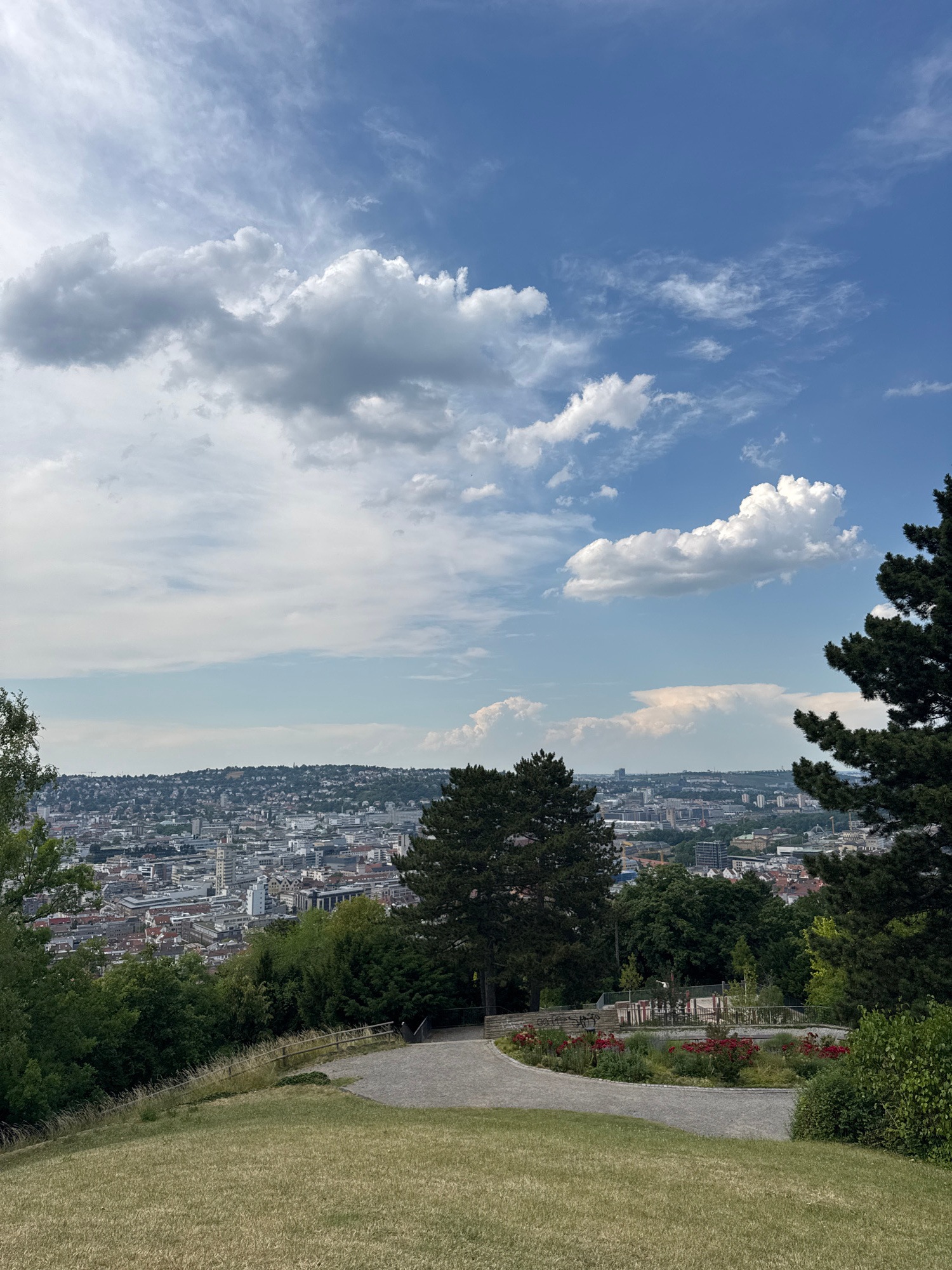 The image captures a panoramic view of Stuttgart, Germany, from an elevated vantage point. The scene is divided between a vast, blue sky filled with fluffy white and wispy clouds, and the sprawling cityscape below. To the right, a well-maintained park area is visible, featuring a gravel path winding through grassy slopes and lush green trees. Red flowers are planted along a section of the path, adding a touch of color. Dense foliage from trees frames the top and left sides of the image, suggesting a location within a park or on a hillside overlooking the city. The cityscape is a dense collection of buildings, with rooftops and facades of varying architectural styles, indicating a densely populated urban area. The time of day appears to be daytime, with bright sunlight illuminating the scene and casting shadows. The weather seems pleasant, with a clear sky and scattered clouds. No people or specific objects are clearly discernible within the image, focusing instead on the landscape and cityscape. There is no visible text in the image.