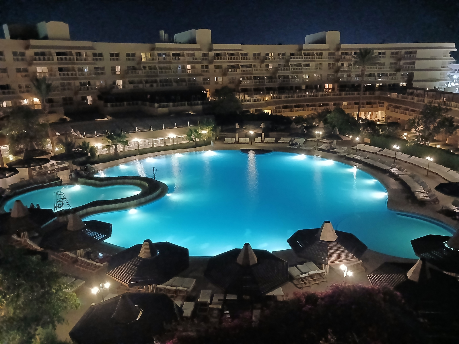 An elevated, wide-angle night view shows a resort complex with a large, illuminated, kidney-shaped swimming pool in the foreground. The pool's water is a vibrant turquoise, with several light sources creating bright spots on its surface. Around the pool are numerous lounge chairs, many of them empty, and several thatched-roof cabanas, some with seating underneath. In the background, a multi-story hotel building with numerous lit windows dominates the scene. Palm trees and other tropical foliage are visible between the pool and the hotel, interspersed with outdoor lighting. The overall atmosphere is serene and illuminated, suggesting a resort environment at night. The location is unknown.