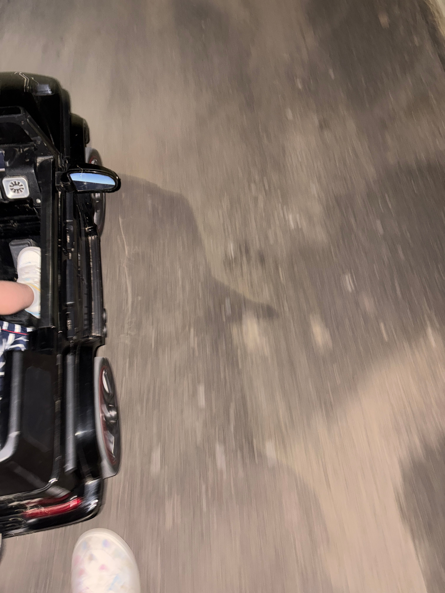 A child's foot in a patterned shoe and a part of their leg wearing striped shorts are visible in the lower left of the image, next to a black toy car. The toy car, resembling a Mercedes-Benz G-Class, is positioned on a grey, textured surface, possibly asphalt. The car is partially obscured, with its side, rear wheel, and a side mirror visible. The image captures a dynamic, slightly blurred perspective, suggesting movement. The lighting is dim, with shadows cast by the car, hinting at an outdoor setting at dusk or night. The background is a blurred expanse of the same grey surface, possibly a street or pathway in Kableshkovo, Bulgaria.