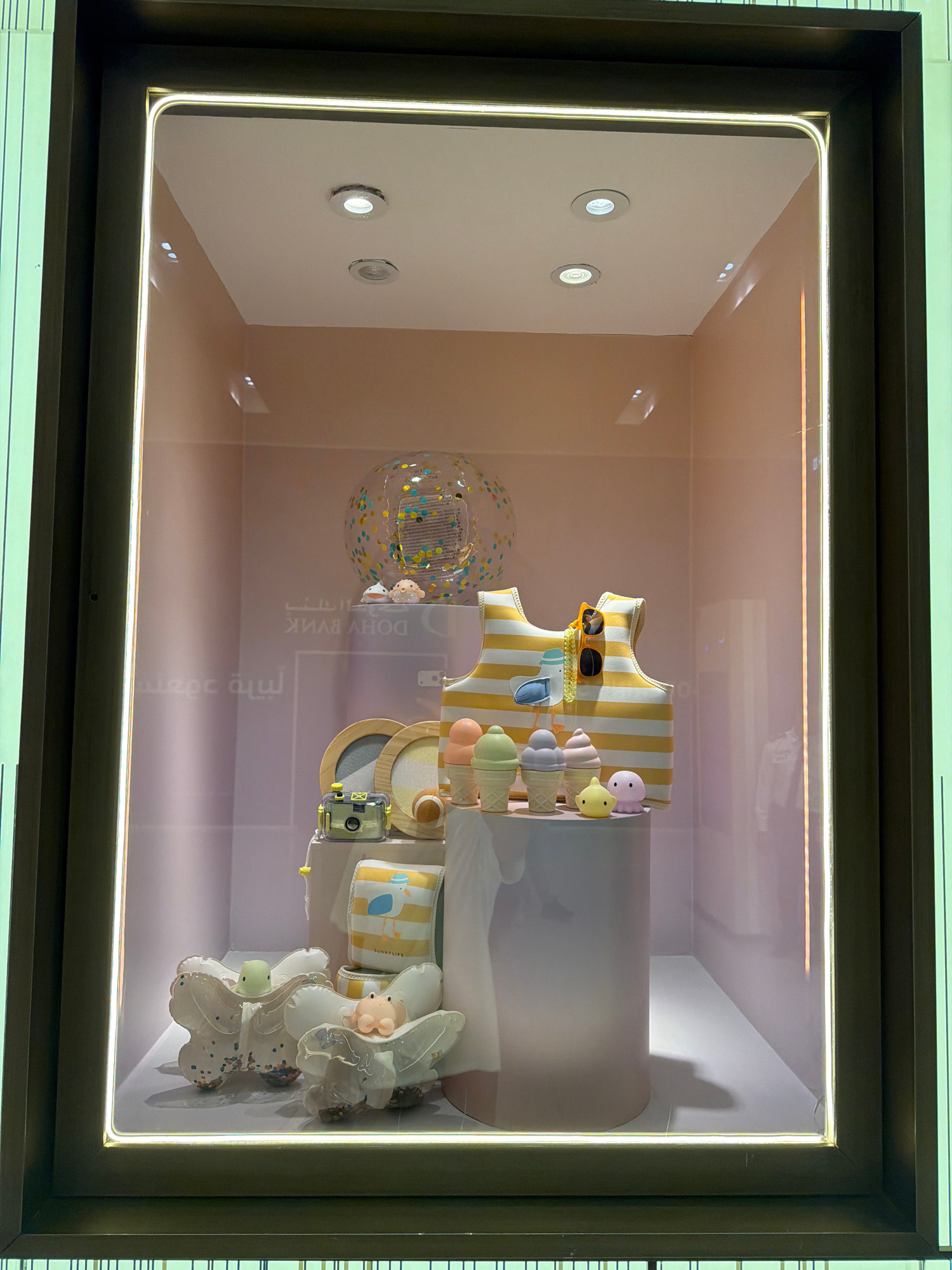 This media file captures a retail display window, likely in a store, showcasing a collection of decorative items. The display features a primarily pastel color palette, dominated by white, yellow, and soft pinks and blues, suggesting a theme related to children's items or a whimsical aesthetic.
Prominently displayed is a yellow and white striped garment, possibly a child's dress or bib, adorned with a pair of orange sunglasses and small accessories that resemble ice cream cones. These cone-shaped objects are colored in various pastel shades and appear to be made of a plastic or ceramic material. Interspersed among these are small, doll-like figures with simple, rounded features and pastel-colored bodies, resembling whimsical characters or toys.
To the left of the central display, a clear, spherical object filled with colorful confetti is visible, adding a festive and playful element. Further to the right, a white, abstract sculpture-like object with flowing lines and embedded confetti can be seen, adorned with similar small, pastel-colored figures. A small camera with a yellow casing is also positioned within the display.
The background of the display is a softly lit, neutral-toned wall, creating a clean backdrop for the colorful items. The overall impression is one of a cheerful, lighthearted, and perhaps summery theme. There is some visible text in Arabic script on a translucent surface within the display, which is partially obscured and difficult to decipher clearly. The lighting within the display is bright and even, with subtle highlights on the objects, suggesting an indoor setting during daylight hours or well-lit retail hours.