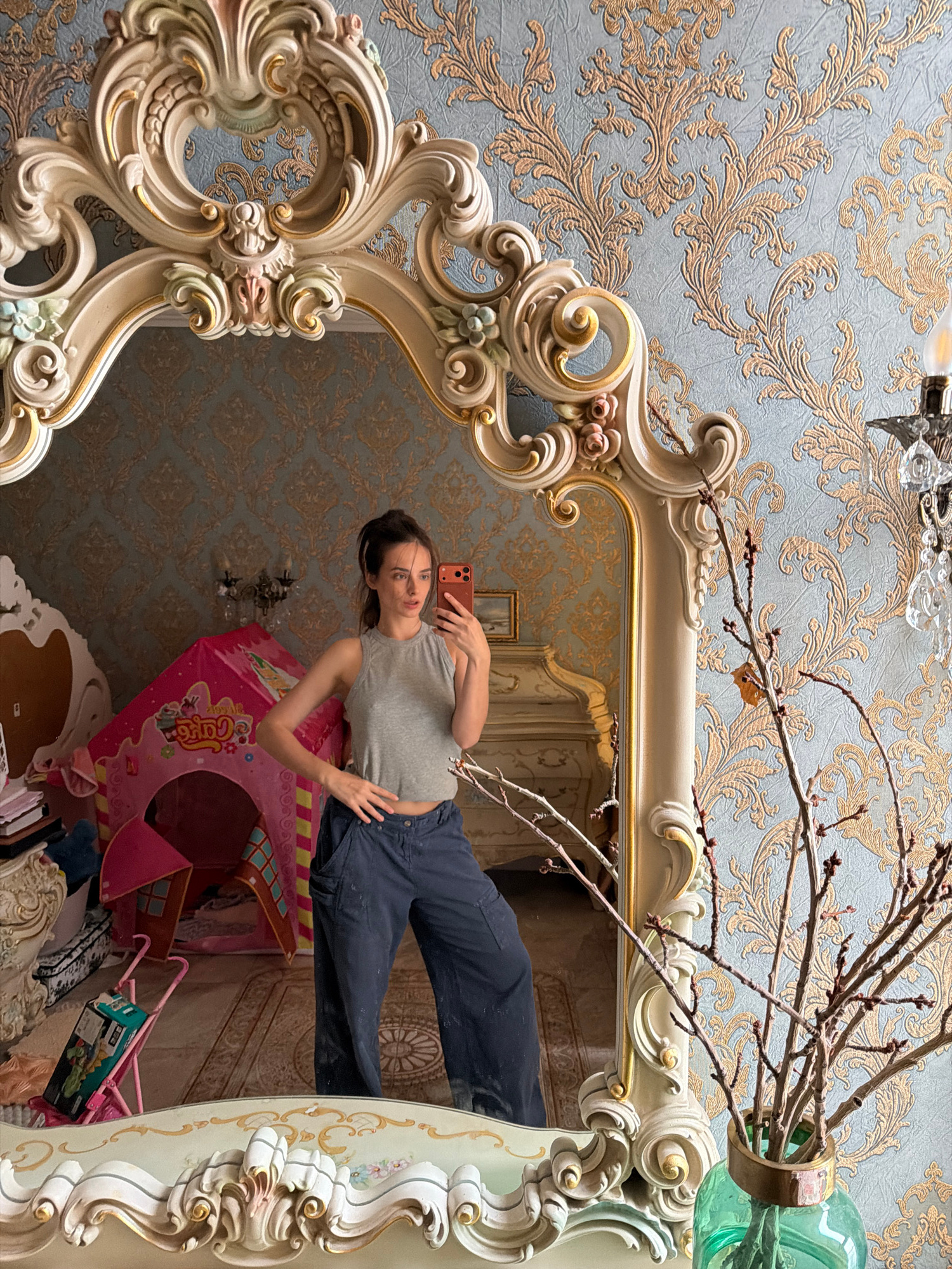 The image shows a young woman taking a selfie in a mirror. She is wearing a grey crop top and dark blue baggy pants. The mirror has an ornate, gilded frame with carved details. In the reflection of the mirror, a pink children's play tent, a stroller, and various other objects are visible, suggesting a residential setting, possibly a bedroom. The wallpaper behind the mirror is ornate and light blue with a gold pattern. A vase with dried branches is in the foreground on the right side of the image. There is no information in the image to suggest this location is in the city of Ufa, Russia.