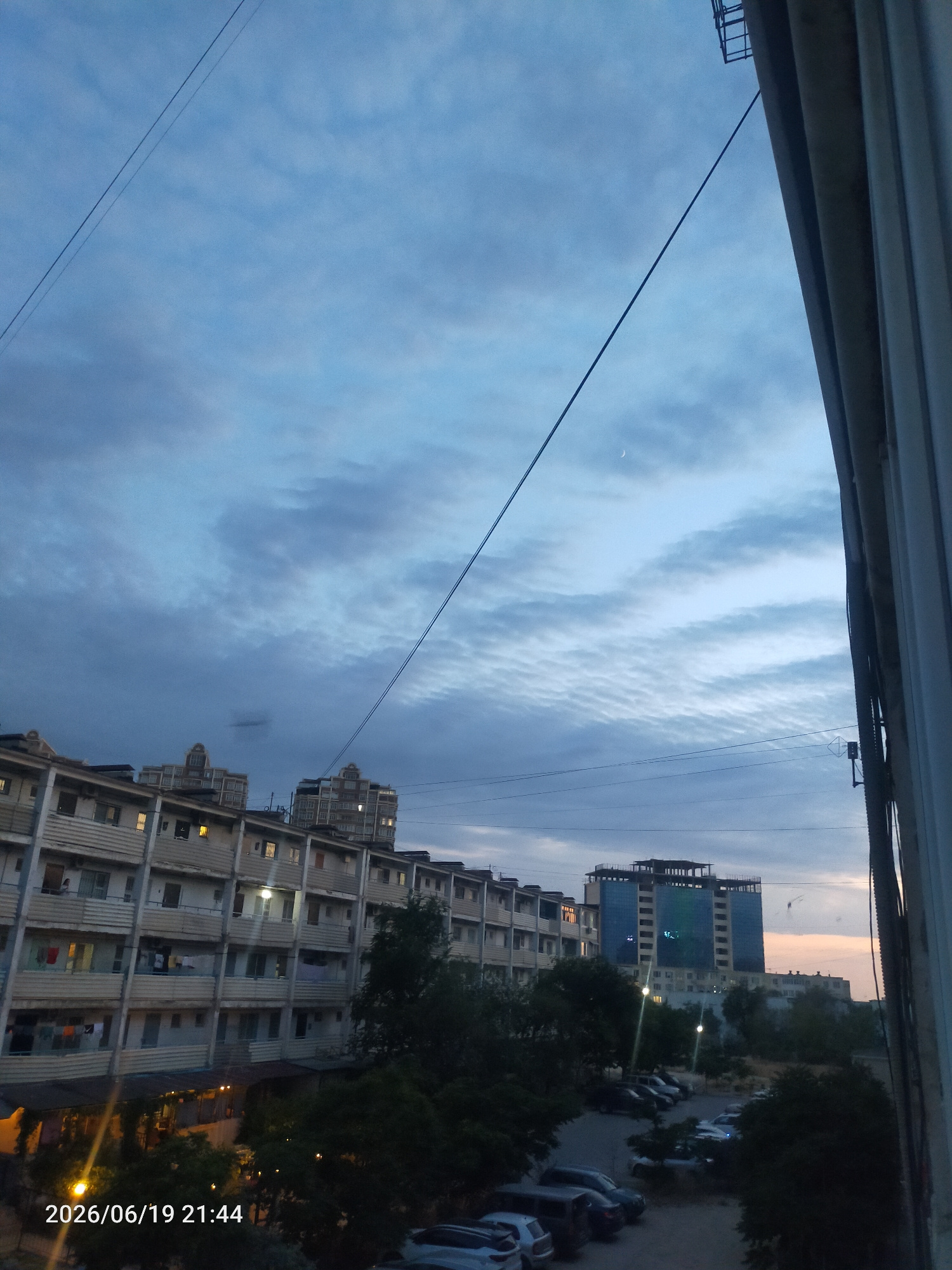 The image captures a twilight scene in an urban environment, likely from a window or balcony, overlooking a residential complex and distant buildings. The sky is a mix of deep blue and lighter gray clouds, with a hint of a crescent moon visible in the upper left quadrant. A power line runs diagonally across the sky from the upper left to the lower right.
In the foreground, a portion of a light-colored, multi-story building with balconies is visible on the left. Laundry is hanging on some of the balconies, and lights are on in several windows, suggesting that it is evening. Below this building, there are trees and some structures, possibly awnings, illuminated by artificial light. A parking lot with several cars is visible further back.
In the middle ground, a row of apartment buildings stretches across the frame. Behind these, a modern, tall building with a glass facade, displaying illuminated rectangular patterns, stands out against the sky. There are also other, less distinct buildings in the distance.
The overall impression is of a residential area at dusk, with a blend of older and newer architecture, and the everyday signs of human habitation. The date and time stamp "2026/06/19 21:44" is present in the lower left corner.