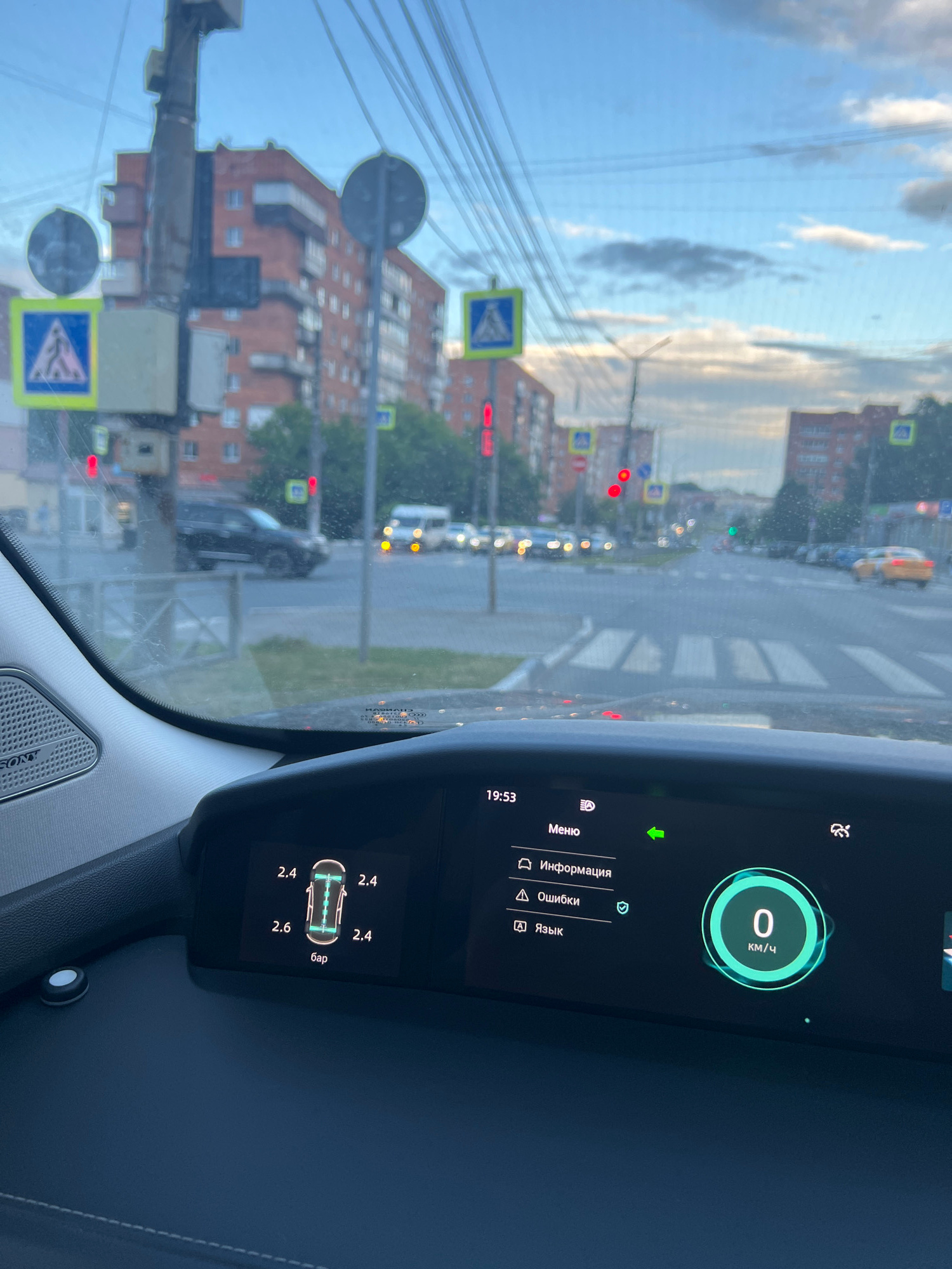 The image captures a view from inside a car, looking through the windshield at a street scene in Tula, Russia, likely during the late afternoon or early evening given the lighting. The dashboard of the car is prominently featured in the foreground, displaying various information. On the left side of the dashboard, tire pressure readings are visible: 2.4 bar for three tires and 2.6 bar for the fourth. The central part of the dashboard shows a menu with options such as "Menu," "Information," "Errors," and "Language." A green arrow indicates a selected option, and a circular display shows "0 KM/H," suggesting the vehicle is stationary.
The exterior view through the windshield shows a street with buildings, traffic lights, and other vehicles. Several residential buildings, characterized by brick facades and multiple windows, line the street. Pedestrian crossing signs with a blue background and a white pedestrian symbol are visible at intersections. Traffic lights are red, indicating vehicles are stopped. A yellow taxi is visible further down the street. The sky is partly cloudy with a soft, diffused light. Overhead, a dense network of power lines crosses the frame, a common sight in Russian urban environments. The overall impression is a typical urban street in Russia, captured from the perspective of a driver.