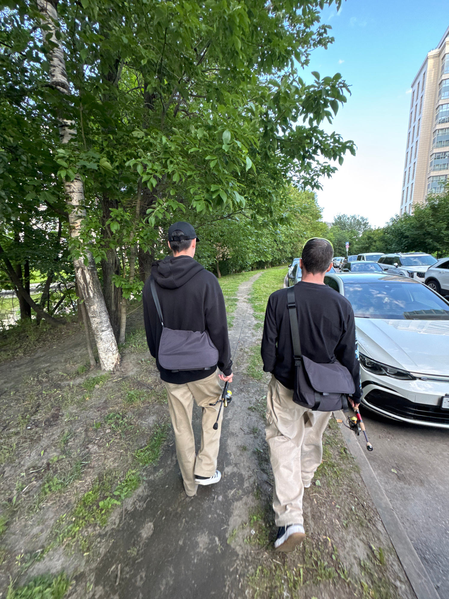 Two males are walking away from the camera on a paved path, which is bordered by a grassy area with trees on the left and cars parked on the right. The male on the left is wearing a black hooded sweatshirt, khaki pants, and a dark gray crossbody bag. He is holding a fishing rod in his right hand. The male on the right is wearing a black long-sleeved shirt, khaki pants, and a dark gray crossbody bag. He is holding a fishing rod in his right hand. In the background, there is a multi-story building with windows and a bright blue sky with a few clouds. The environment appears to be an urban setting in Saint Petersburg, Russia, possibly a park or residential area.