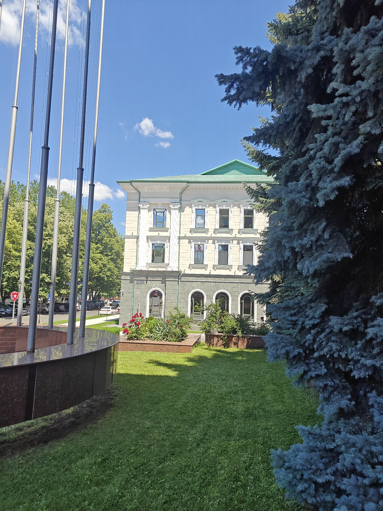 This image captures a daytime scene featuring a multi-story building with a green roof, located in the city of Chișinău, Moldova. The building exhibits classical architectural elements, including columns and arched windows on the lower level. In the foreground, a large, dark blue spruce tree partially obscures the view of the building. To the left, a series of tall, silver poles are visible, likely part of a monument or flagpole installation, alongside a curved, dark-colored structure that appears to be made of polished stone, possibly a fountain or memorial base. A well-maintained green lawn stretches across the mid-ground, with a flowerbed featuring red roses situated in front of the building. The background reveals a street with parked cars and lush green trees lining the area. The sky is bright blue with a few scattered white clouds, indicating clear weather conditions. There are no people visible in the image.