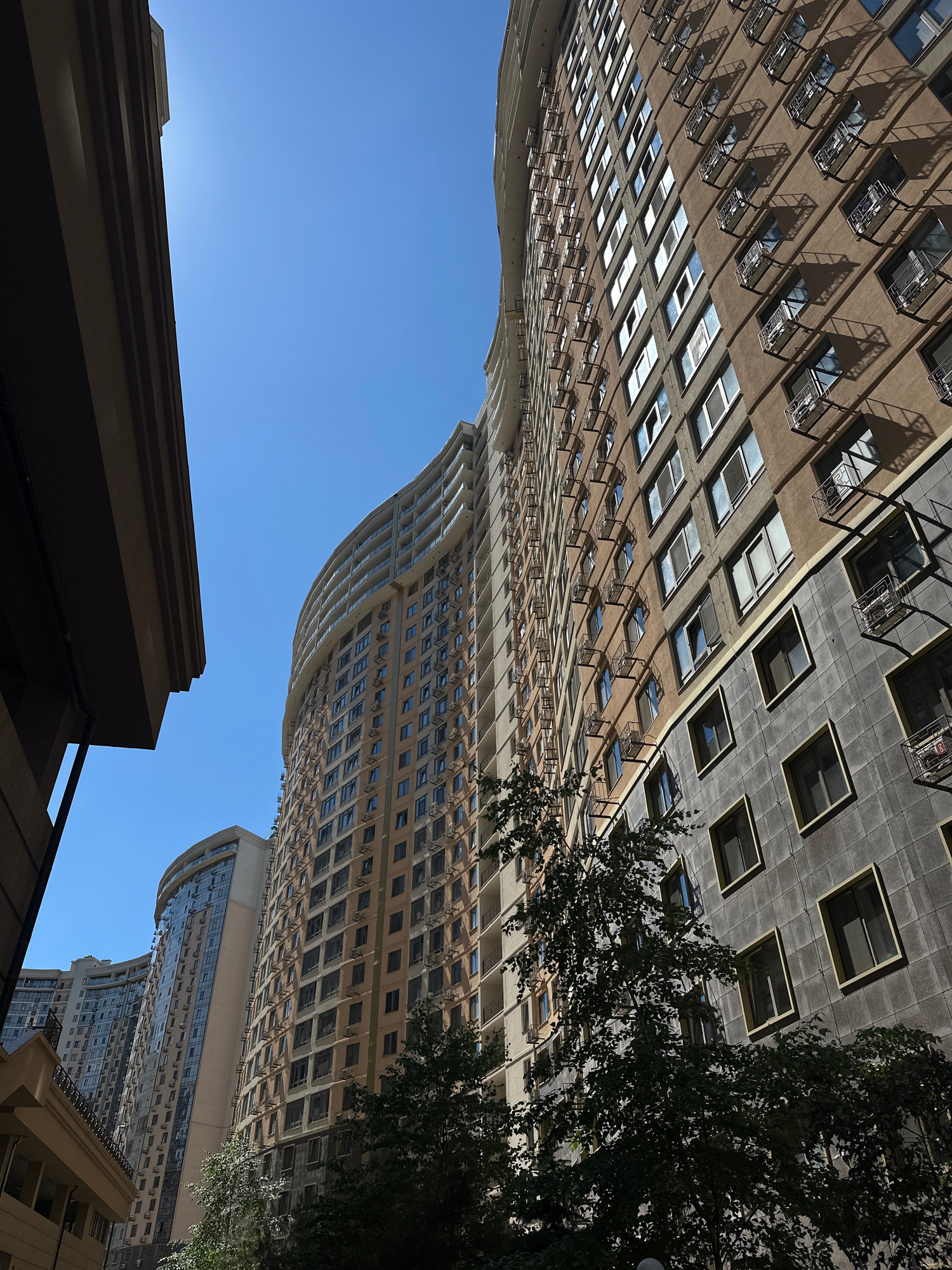 In Odesa, Ukraine, a low-angle shot captures a perspective looking up at the facades of several contemporary residential high-rise buildings against a clear blue sky. The building on the right, a prominent feature, is characterized by its curved architecture and a textured exterior in shades of beige and grey. Numerous windows punctuate the facade, many equipped with external air conditioning units and decorative metal grates. The building on the left, partially visible, is also a modern structure with a lighter facade and extensive glazing. In the foreground, the edge of another building, darker in color and with horizontal banding, partially obscures the view on the left. Trees with dense green foliage are situated at the base of the buildings, casting shadows on the lower portions of the structures. The overall scene depicts urban architecture under bright daylight conditions.
