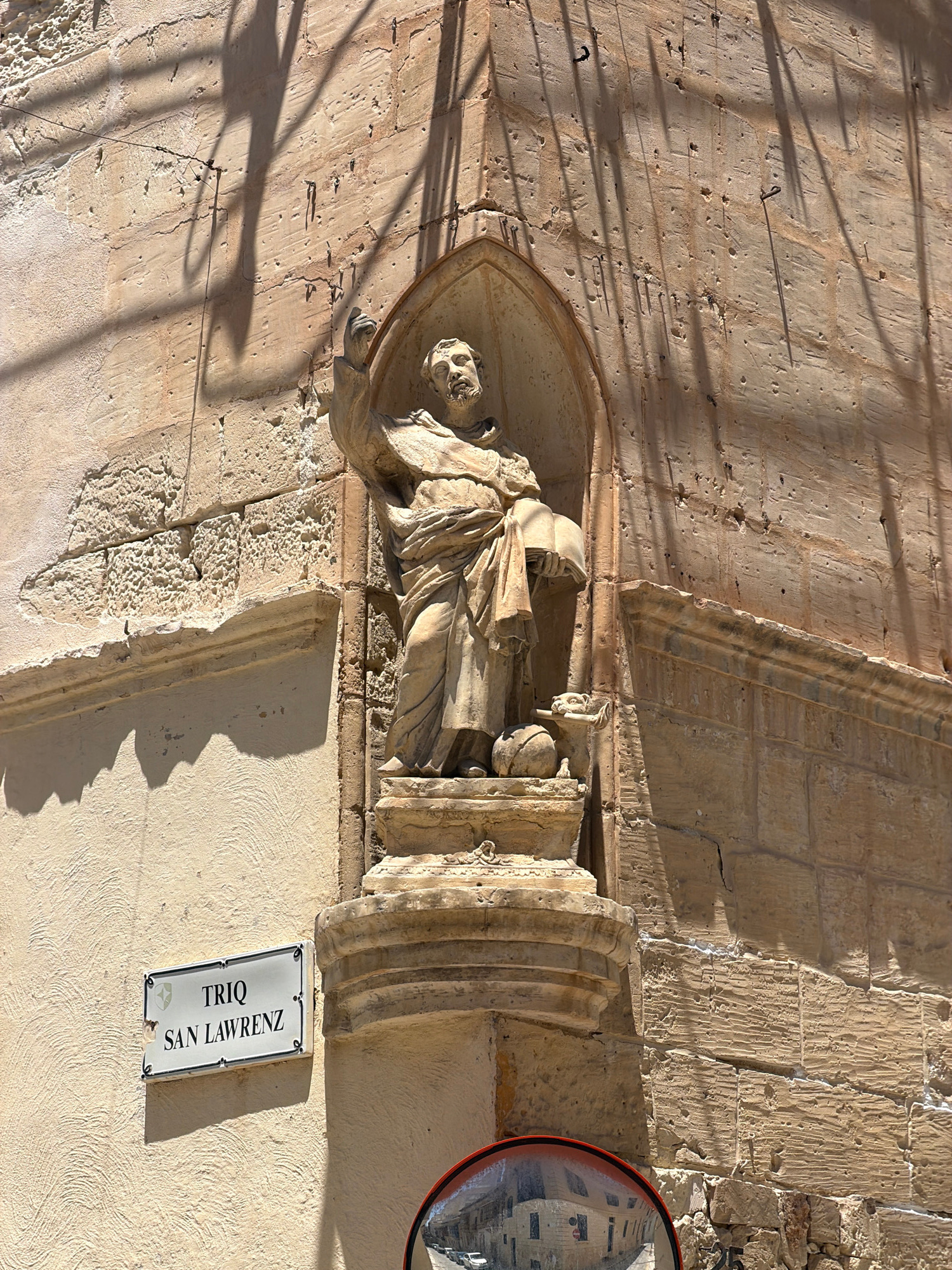 This image shows a stone niche on a building, possibly an older structure, with a sculpture of a person. The sculpture appears to be a religious figure, depicted in a robe, with their head bowed and hands in a prayerful or supplicating gesture. The person in the sculpture seems to be cradling something or offering it. The niche is arched and adorned with decorative stone carvings.
The wall surrounding the niche is made of rough-hewn stone blocks, some of which show signs of wear and tear. Shadows cast by a grate or window are visible across the wall, indicating a sunny day. The light suggests it is daytime, likely midday or afternoon given the angle of the shadows.
On the wall below the niche, there is a rectangular street sign with a white background and black text. The text reads "TRIQ SAN LAWRENZ", with a small logo of a Maltese cross to the left of the text. "Triq" means "street" in Maltese, and "San Lawrenz" translates to "Saint Lawrence". This text provides a strong clue to the location being Malta, specifically a street named after Saint Lawrence.
In the upper right corner, a portion of a convex mirror is visible, reflecting the surrounding architecture, which appears to be of stone construction and likely in a Mediterranean style. The overall impression is one of historical architecture bathed in sunlight.