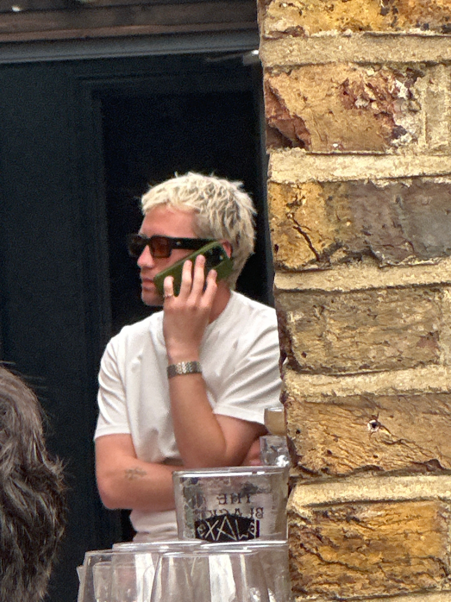 A person with bleached blonde hair and sunglasses is talking on a cell phone, holding it to their right ear with their right hand. They are wearing a white t-shirt and a watch on their left wrist. A tattoo is visible on their left forearm. The person is standing next to a brick wall and appears to be in an outdoor setting. In the foreground, there are several clear glasses. The background is dark, suggesting an indoor or shaded area. The brick wall is textured with visible mortar lines and variations in brick color.