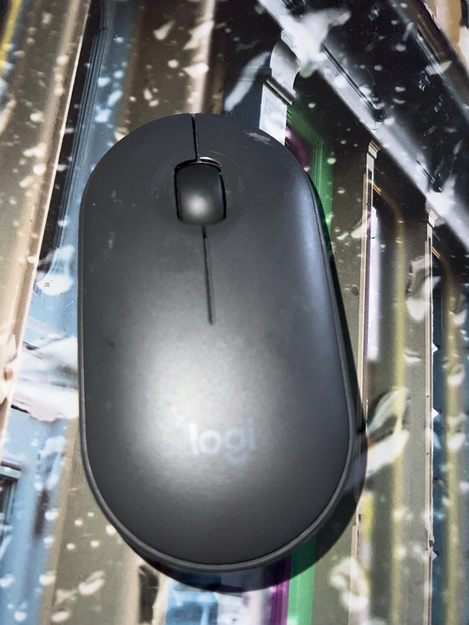 The image shows a dark gray Logitech computer mouse positioned on a patterned surface. The mouse is the primary subject, with its curved top and a single button visible in the foreground. The button appears to be black and slightly raised. A thin line runs horizontally across the mouse's surface, likely separating the left and right click functions. The Logitech logo is subtly embossed on the upper right portion of the mouse, appearing as a lighter gray imprint. The background is a busy, abstract pattern with streaks of black, white, and hints of green and purple. The lighting creates a strong shadow on the right side of the mouse and highlights the curved surface. The overall impression is that of a close-up shot of a computer accessory, possibly on a desk or a specialized mousepad, with the focus sharply on the mouse itself. There are no people or specific environmental details like weather or time of day that can be discerned from the image. The location context provided suggests this image was taken in Novosibirsk, Russia, but there are no visual cues within the image to confirm this.