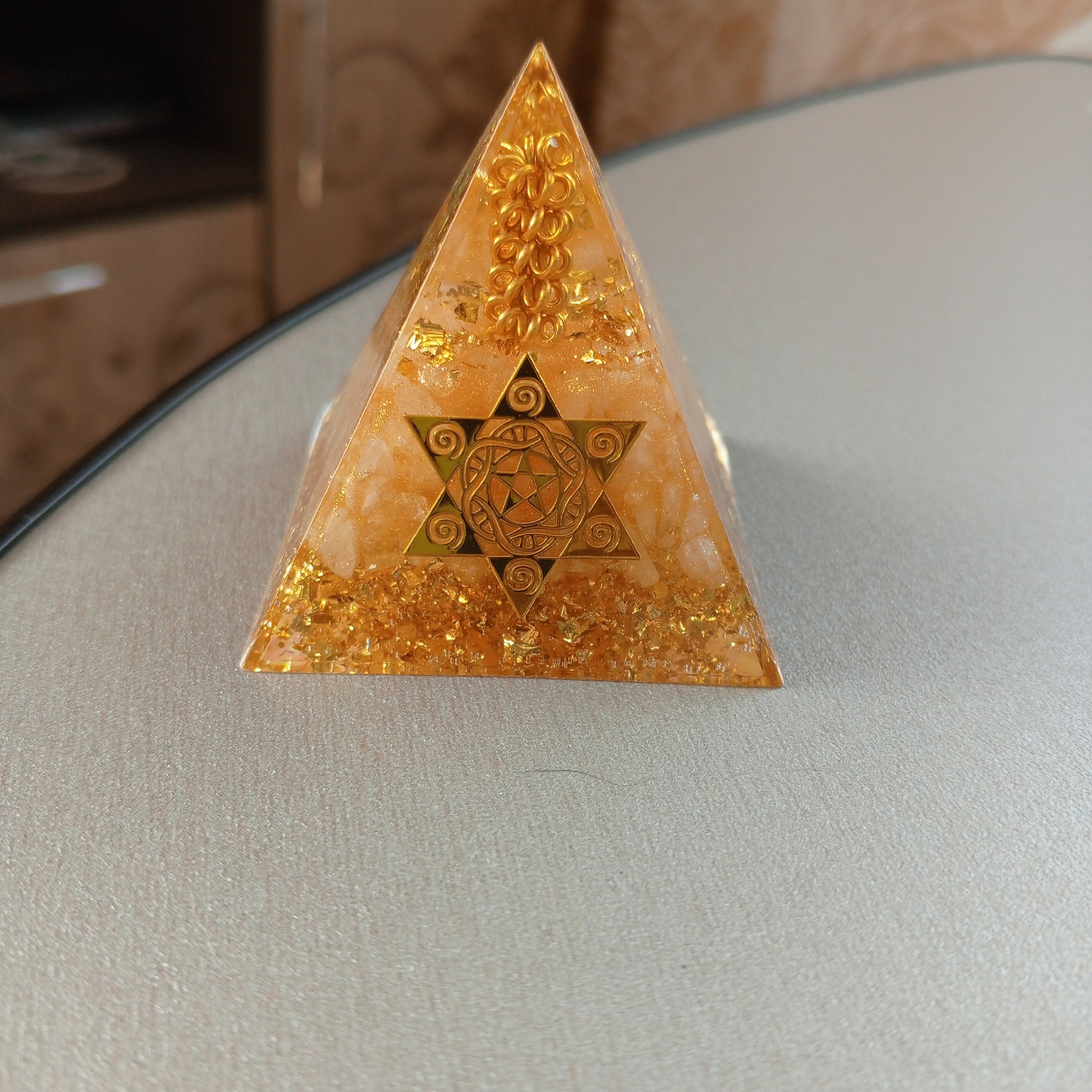 A golden pyramid sits on a light gray surface, possibly a table. The pyramid has a star-shaped symbol embedded in its center, with intricate spiral designs within the star. The pyramid itself appears to be made of a translucent material, possibly resin, with flecks of gold leaf or glitter suspended within it, giving it a shimmering, ethereal appearance. The background is blurred, showing hints of a domestic setting with warm-toned furniture. The overall lighting is soft, casting a subtle shadow from the pyramid onto the surface. The location of Шелопугино, Russia is not depicted in this image.