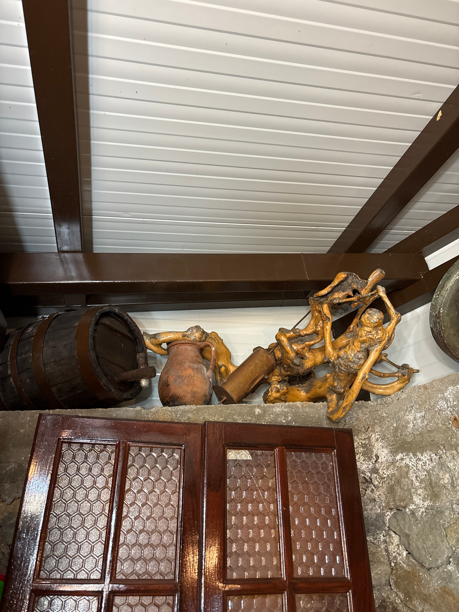 This image depicts a rustic interior setting with an overhead white paneled ceiling supported by dark brown beams. On a rough concrete shelf below, there are several objects. To the left is a dark wooden barrel with metal bands. Next to it is a small, reddish-brown ceramic jug with a handle. To the right of the jug is a piece of gnarled, light-colored wood carved into figures that appear to be interacting. In the foreground, two dark, polished wooden doors with textured glass panels are visible. The overall scene suggests a storage area or a display in a traditional or historical setting, possibly in the city of Zlatograd, Bulgaria, known for its preserved ethnographic architecture.