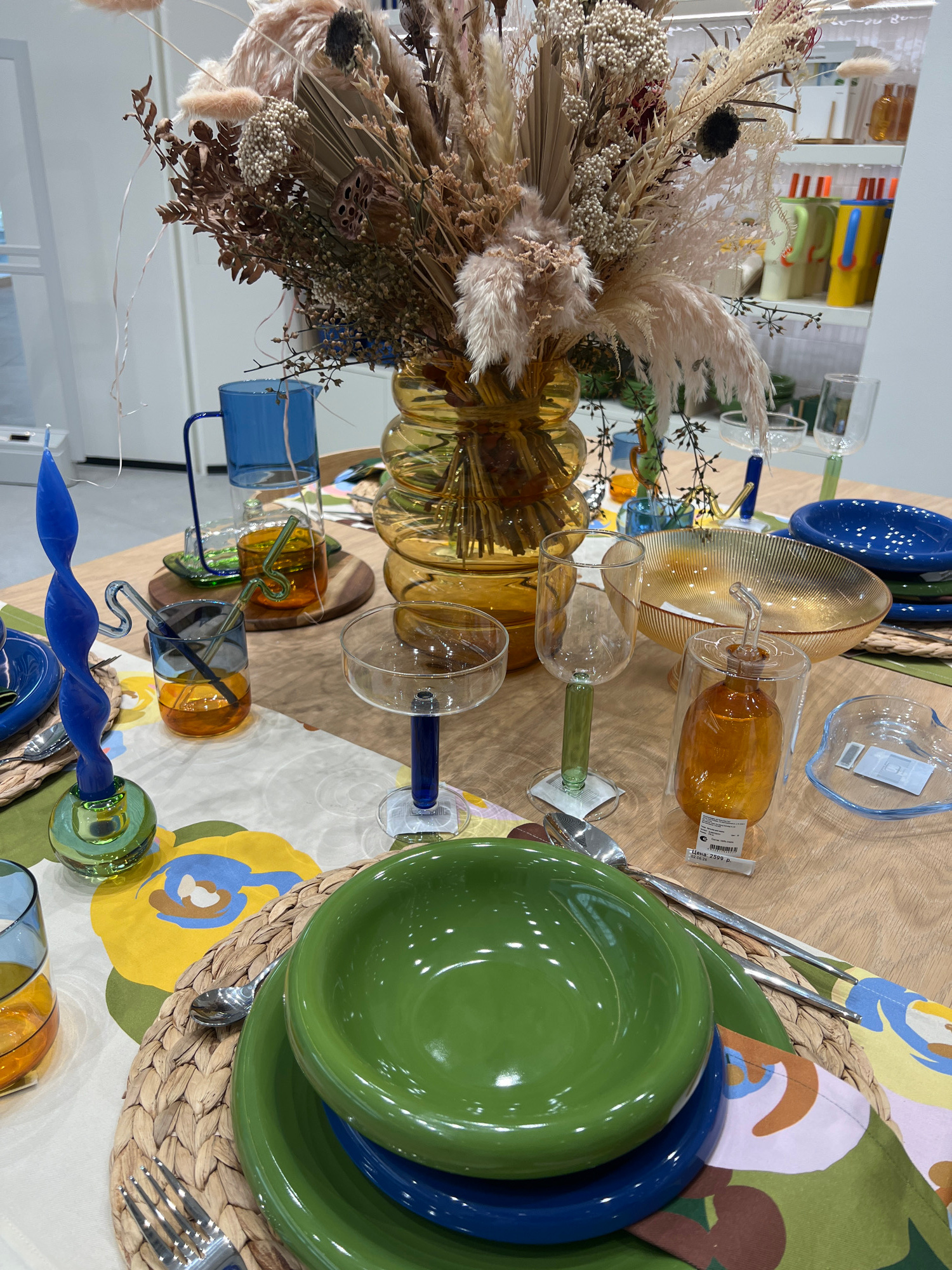 The image depicts a table setting with a floral-patterned tablecloth. A large bouquet of dried flowers in a ribbed amber vase is the centerpiece. Several glass items are arranged on the table, including blue and amber tumblers, champagne coupes with colored stems, and a glass cruet. There are also plates and bowls in blue and green, along with cutlery. A twisted blue candle in a green glass holder is visible on the left. In the background, shelves display various colored pitchers and bottles. The scene is indoors, likely in a retail setting given the shelving unit and product displays. There is no indication of the specific location in Saint Petersburg, Russia.