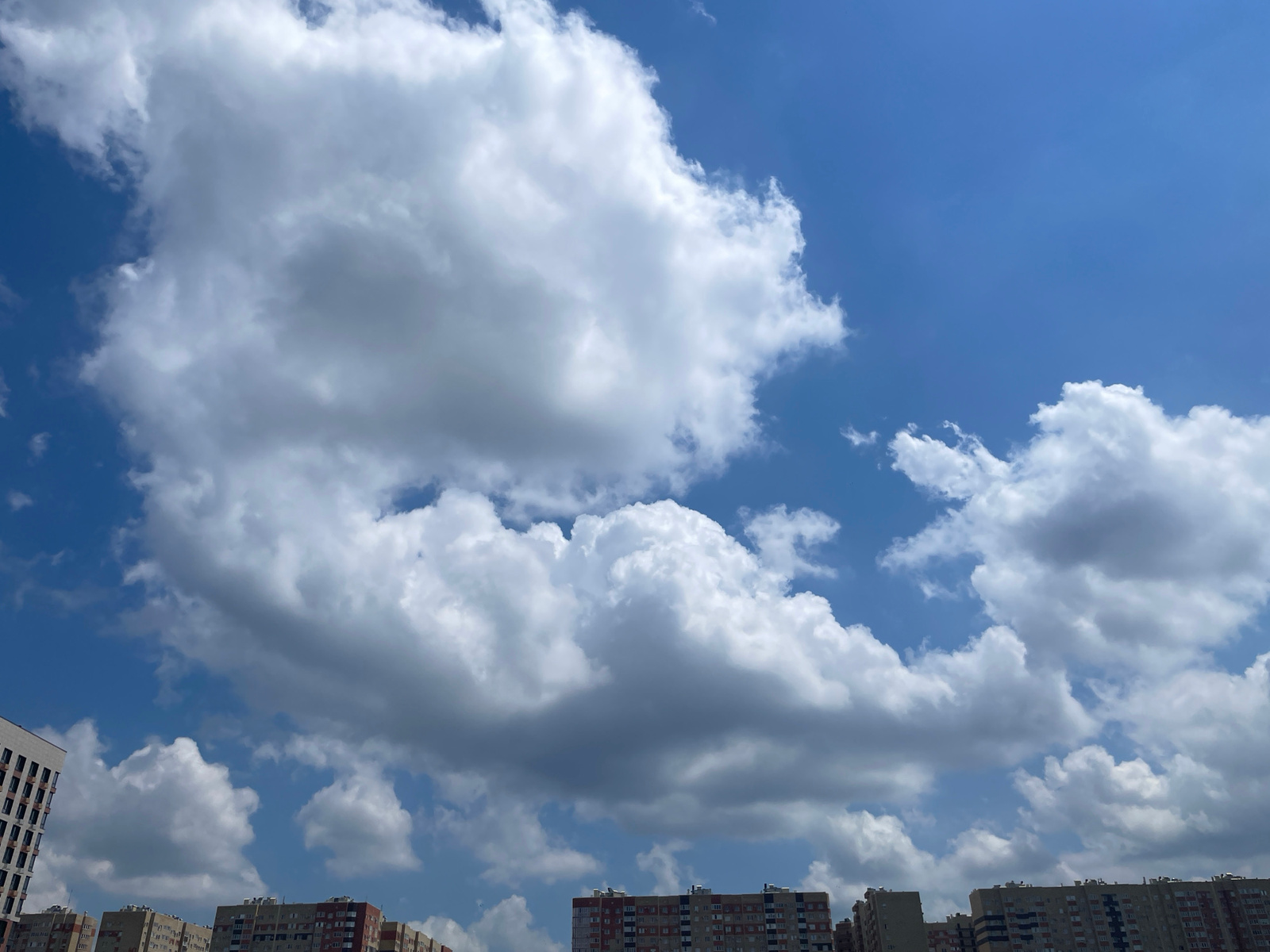A wide-angle, eye-level shot captures a bright blue sky filled with dramatic, billowing white and grey clouds. The lower third of the image is dominated by the rooftops of several multi-story apartment buildings. The buildings are uniform in height and appearance, suggesting a residential development. The architecture is modern, with visible windows and balconies. One building on the far left is taller than the others and has a distinct, lighter colored facade. There are no people or vehicles visible in the image. The scene depicts a clear day over a city, likely a residential area in Stavropol, Russia, given the building styles and sky.