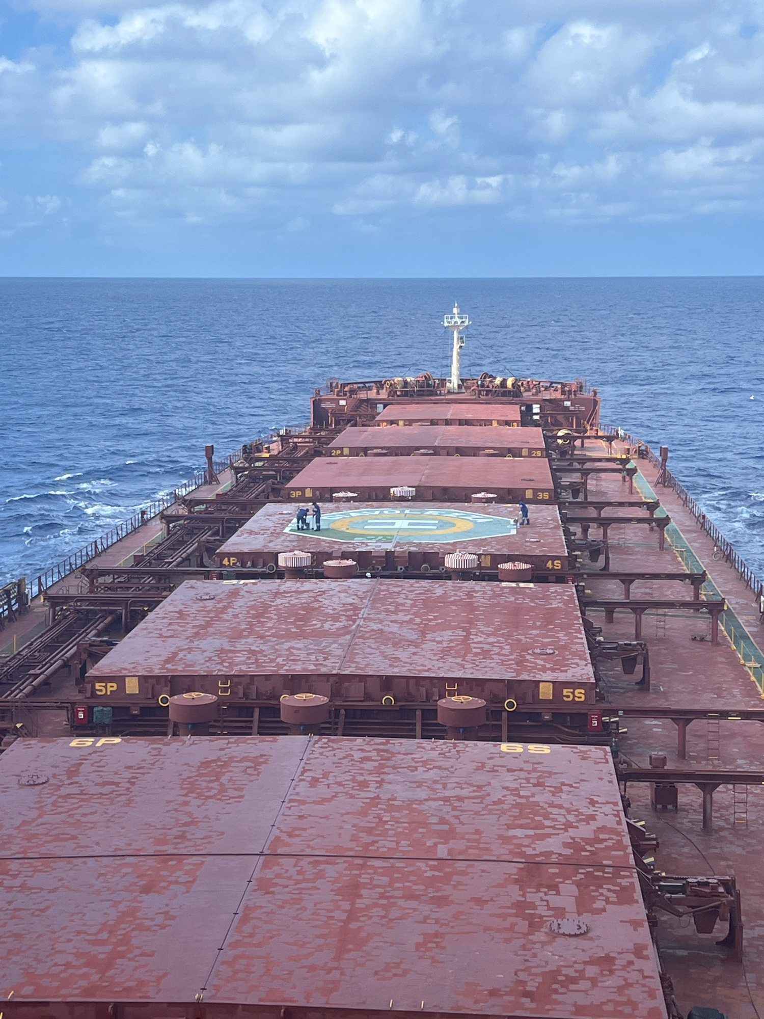 This is a medium-shot, elevated, eye-level, horizontal, outdoor, daytime, high-angle, full shot of the deck of a large cargo ship sailing on a blue ocean under a cloudy sky. The ship's deck is covered with red hatches and metal structures. A helipad with a yellow and blue marking is visible in the middle of the deck. Several people are present on the deck, some gathered on the helipad and others standing further back. The sea is calm with small waves. The image is taken from the stern of the ship, looking forward.