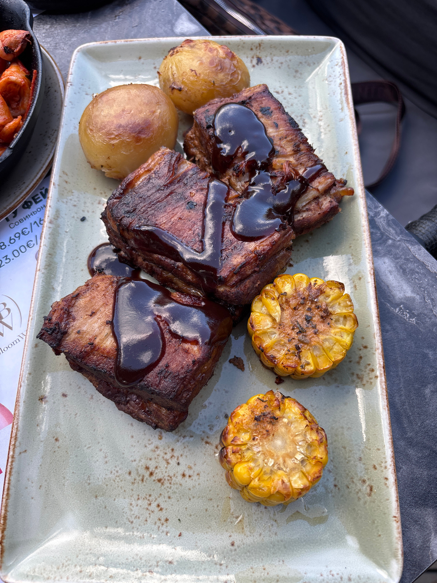A close-up, overhead shot shows a rectangular ceramic plate with a light gray speckled glaze and a brown rim. On the plate are three pieces of barbecued pork ribs, generously drizzled with a dark, glossy sauce. The ribs have a dark, caramelized exterior and appear to be tender. Accompanying the ribs are two small, golden-brown roasted potatoes and two round slices of grilled corn on the cob, with visible char marks and kernels. To the left of the plate, a portion of a black cast-iron dish is visible, containing what appears to be cooked sausage or meat. A white paper menu with Cyrillic text and a logo is also partially visible on the left side of the frame. The plate sits on a dark, textured surface, possibly a table, with a blurred background that suggests an outdoor setting. The overall scene depicts a prepared meal, likely at a restaurant. The menu text includes prices like "8.69€/16" and "23.00€/", hinting at a European location. Considering the context and style of food, this could be a scene from a restaurant in Nesebar, Bulgaria, known for its historical sites and culinary offerings.