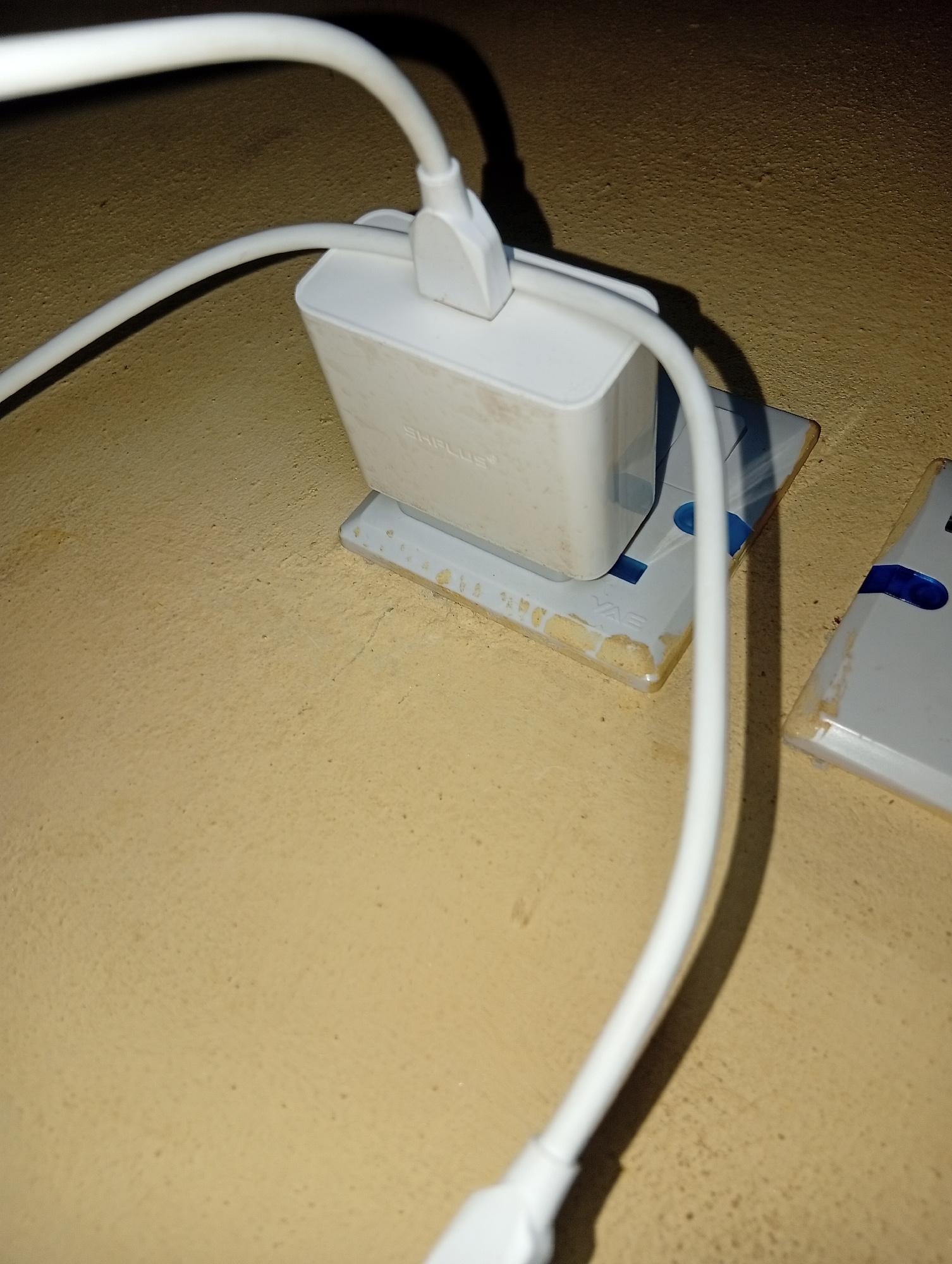 A white power adapter is plugged into a beige wall socket. Two white cables extend from the adapter, one curving upwards and out of frame, and the other curving downwards. The wall socket is partially obscured by the adapter and cables. The wall surrounding the socket is a textured, light brown color. Another similar wall socket is visible to the right of the primary one.