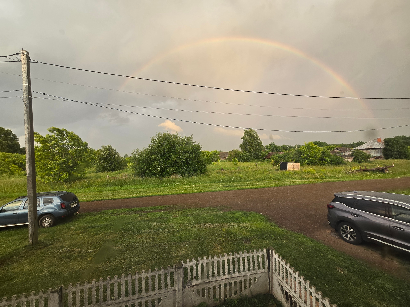 This image depicts a rural landscape under a cloudy sky, with a rainbow arching across the horizon. In the foreground, a white picket fence encloses a patch of green grass. To the left, a blue SUV is parked on the grass near a utility pole. A dirt path runs horizontally across the midground, bordered by more grass and lush green trees and bushes. In the distance, several buildings, including houses and what appear to be agricultural structures, are visible, hinting at a settlement. The overall scene suggests a post-rain, possibly in the city of Jelpachiha, Russia, given the architectural styles and vegetation. No people are visible in the image.