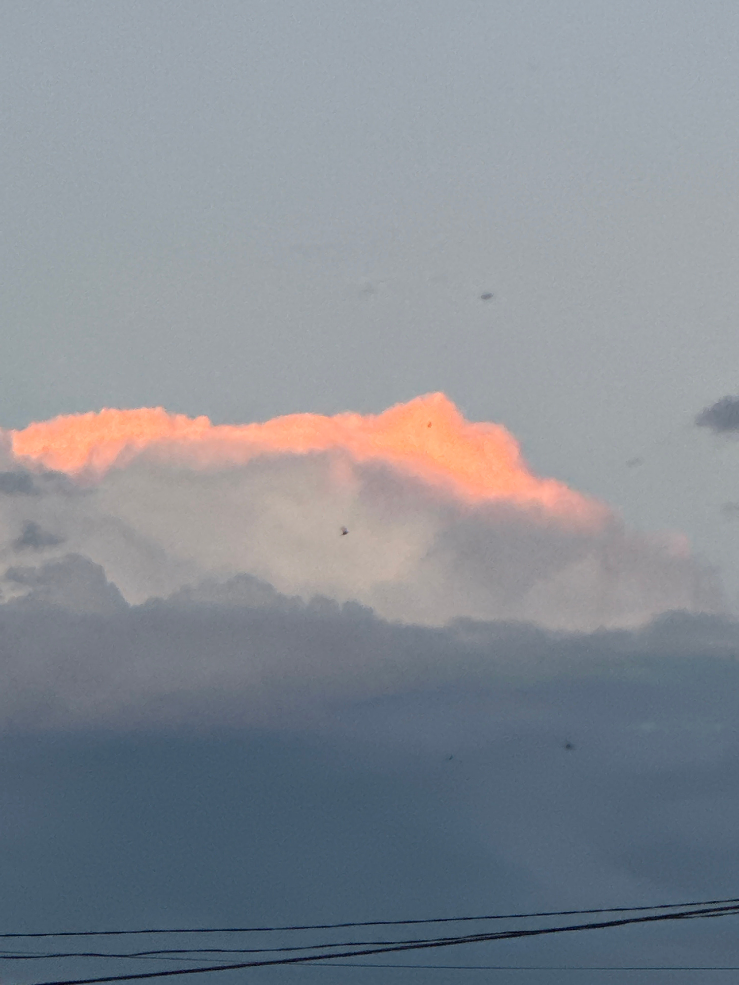 A horizontal composition captures a sky at dusk with multiple layers of clouds. The uppermost clouds are illuminated with a bright orange hue, suggesting the last rays of sunset. Below this, a layer of lighter grey clouds is visible, and the bottom layer consists of darker, heavy grey clouds. A few faint, dark specks are scattered across the sky, possibly distant birds or insects. In the lower portion of the image, three thin, dark horizontal lines represent power or telephone wires. The scene is devoid of human figures or identifiable landmarks within the city of Ufa, Russia.