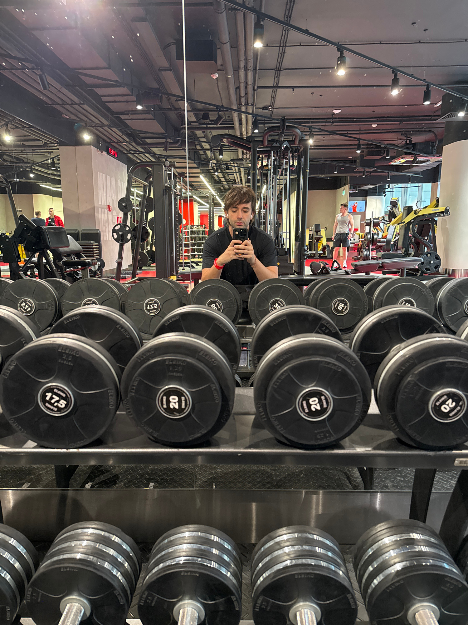 This is a vertically oriented, low-angle, candid photograph taken inside a modern, well-lit gym. The scene is dominated by a rack of black weight plates with visible "ELEIKO" branding and weight markings like "20", "17.5", and "25". To the right, a set of dumbbells with multiple plates can be seen. In the background, various gym equipment, including machines and bars, are visible, along with what appears to be a digital display with red numbers.
In the foreground, a young man with dark hair is taking a selfie with his phone, visible in his hands. He is wearing a black t-shirt and has a red wristband on his left arm. His expression is neutral. Further in the background, another man in a grey tank top and shorts is standing near a piece of equipment, possibly mid-workout. The gym appears to be a commercial establishment, possibly in Moscow, Russia, given the location context. The lighting suggests it's daytime, and the overall atmosphere is that of a typical gym environment focused on strength training. The image captures a moment of self-documentation during a workout session.