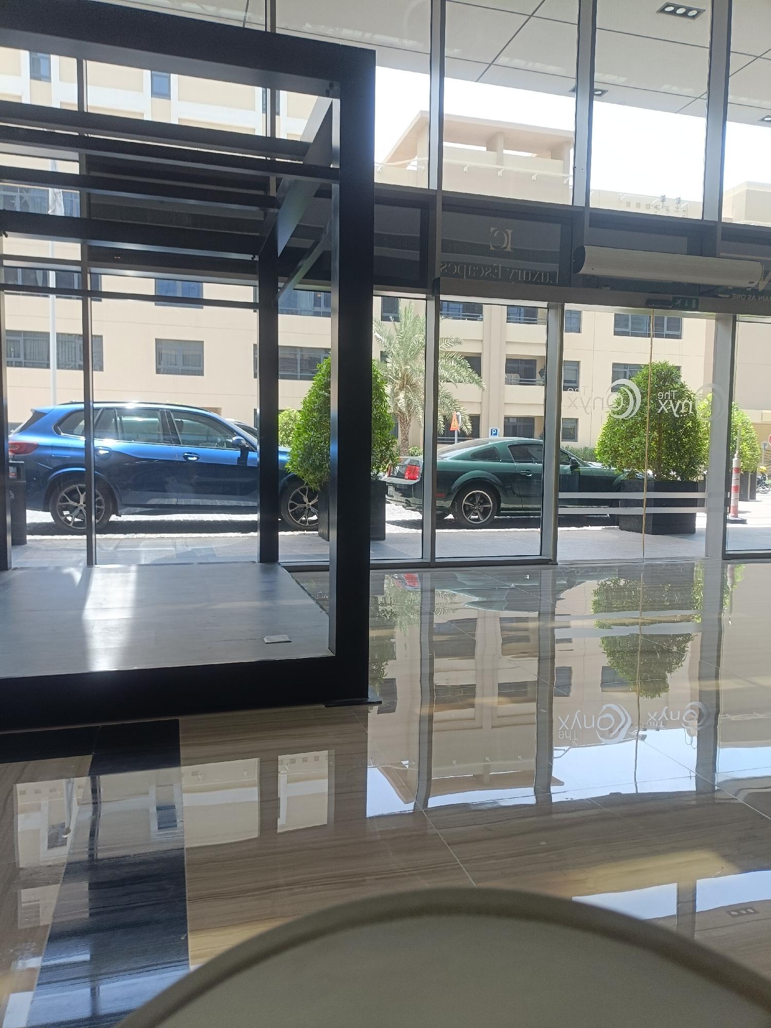 The image depicts a modern architectural interior with a reflective floor, likely a hotel lobby or a commercial building entrance in Dubai, United Arab Emirates. Through large glass windows, two cars are visible parked outside. On the left, a blue SUV is parked. To the right, a dark green coupe is parked next to a planter with lush green foliage. The background reveals a multi-story building with light-colored facade and numerous windows, typical of Dubai's urban landscape. The reflective floor shows inverted reflections of the building and the sky, creating a sense of depth and elegance. The lighting suggests it is daytime, with sunlight creating bright spots on the floor. There are no people visible in the scene. The overall impression is one of sophistication and contemporary design.