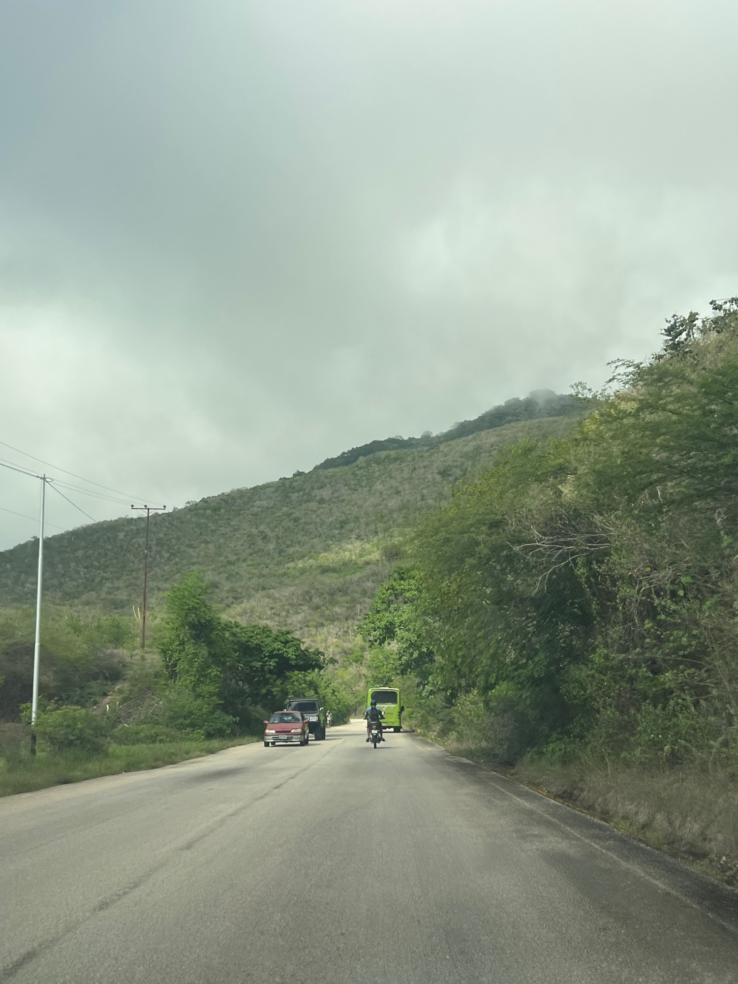 A road curves through a mountainous, sparsely vegetated landscape under a cloudy sky. The road, cracked and faded in places, runs from the foreground towards the horizon. On the left side of the road, a tall utility pole with wires stretches across the frame. Green trees and shrubs line both sides of the road, with a more densely wooded hillside rising on the right. The left side of the scene also features a smaller, sparse hillside with some lighter vegetation.
Vehicles are visible on the road. In the middle distance, a red car and a dark SUV are traveling in the same direction as the viewer. Further ahead, a green bus is also in motion, and a motorcyclist is in front of it. The top of a distant mountain is obscured by mist. The overall impression is a rural, somewhat arid environment.