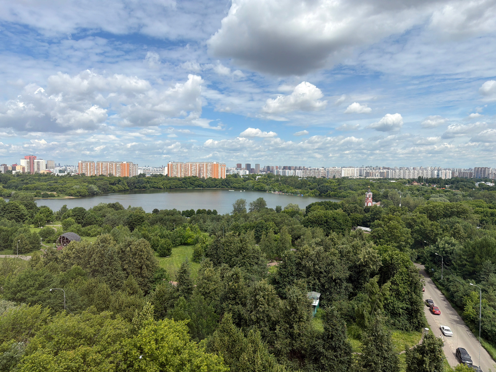 A panoramic, elevated view captures a city skyline beyond a verdant park and a body of water under a cloudy sky. The foreground is dominated by dense, lush green trees. A paved road winds through the trees on the right side, with several cars driving on it: a white sedan, a black sedan, a red car, and another dark-colored car. In the middle ground, a large lake or reservoir is visible, with some structures or boats on its surface. Further back, a park area with grassy clearings and scattered trees sits between the water and the city. A distinctive geodesic dome-like structure is situated within this park. A red-domed church with a white tower stands out amongst the trees near the water's edge. The background is a cityscape featuring numerous apartment buildings, predominantly in orange and white, stretching across the horizon. The sky is a mixture of blue and large, fluffy white and gray clouds.