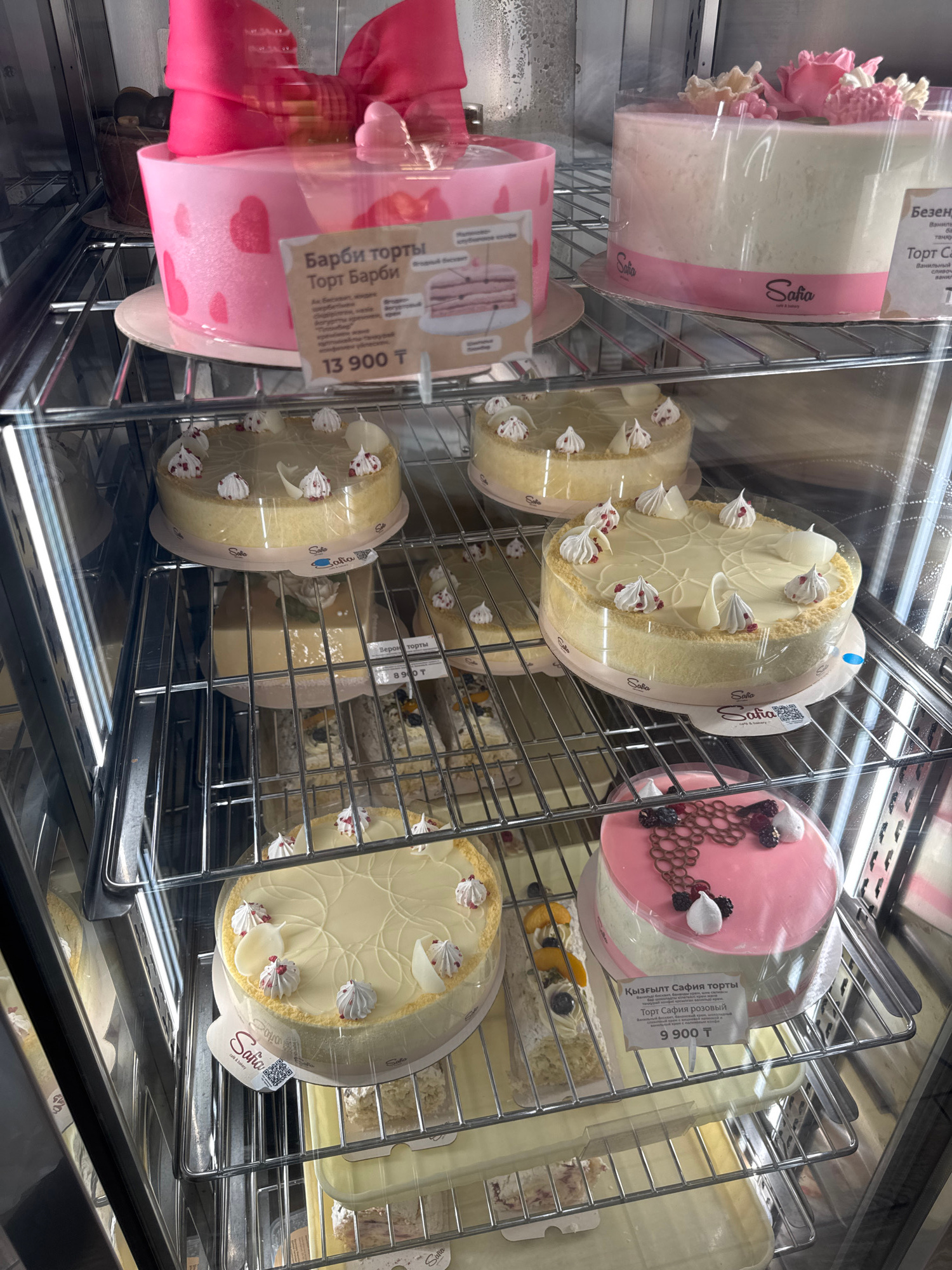 This is a tilted, eye-level shot of cakes displayed in a refrigerated glass case, likely in a bakery or cafe in Almaty, Kazakhstan. The display features multiple round and oval cakes, some with intricate frosting designs, fruit toppings, and decorative elements like chocolate swirls and meringue kisses. Several cakes are packaged in clear plastic containers with branded labels.
Notable text visible on the labels includes "Sofia cafe & bakery," "Торт Барби" (Barbie Cake), "Ягодный бисквит" (Berry Biscuit), "Верона торты" (Verona Cakes), and "Қызғылт Сафия торты" (Pink Safia Cake). Prices are also displayed, with "13 900 T" (likely Tenge, the Kazakh currency) for the Barbie cake, "8900 T" for a Verona cake, and "9 900 T" for the pink Safia cake. The overall scene suggests a retail environment where these cakes are being offered for sale. The lighting appears to be artificial, consistent with an indoor display case. There are no people visible in the image.