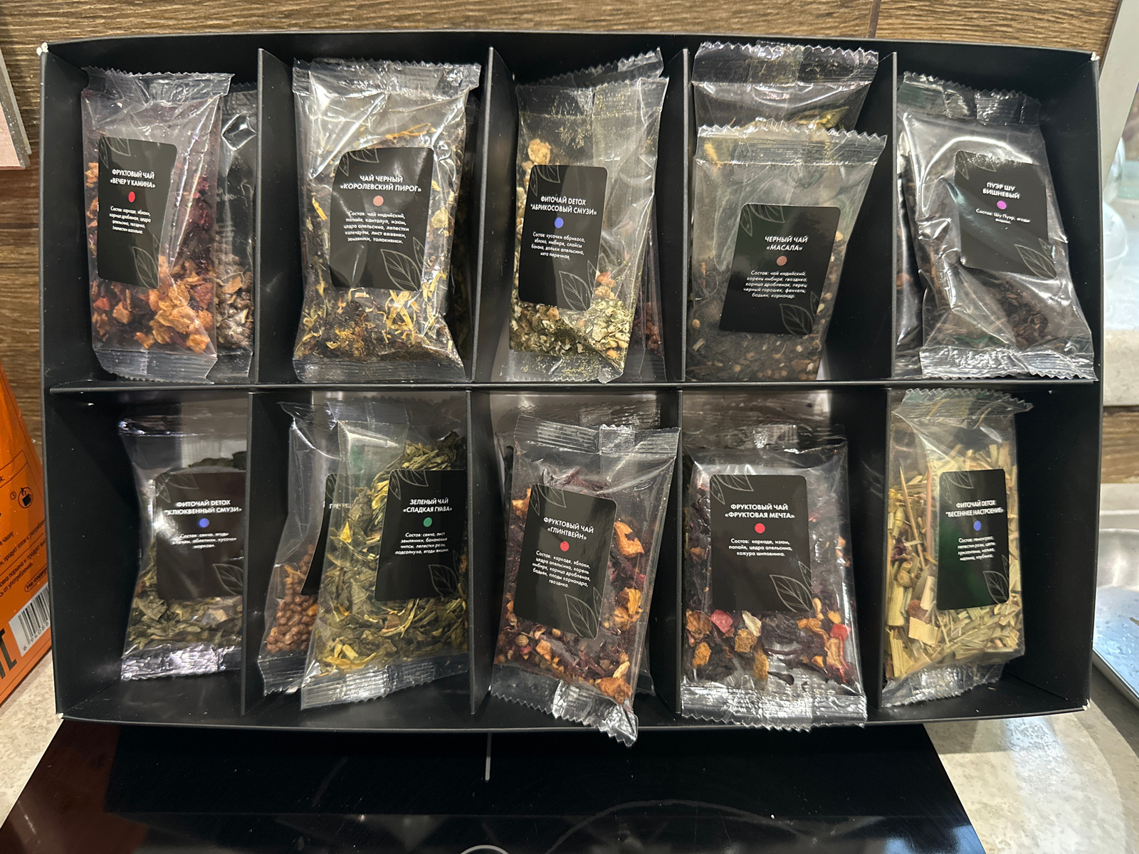This is a top-down, medium shot of a black, rectangular box filled with individually packaged teas. The box is divided into sections, with ten tea packages visible in two rows of five. Each package is a clear plastic pouch with a black label affixed to the front. The labels have text in Cyrillic, indicating the type of tea and its ingredients.
The first row of teas, from left to right, are labeled: "ФРУКТОВЫЙ ЧАЙ «ВЕЧЕР У КАМИНА»" (Fruity Tea "Evening by the Fireplace"), "ЧАЙ ЧЕРНЫЙ «КОРОЛЕВСКИЙ ПИРОГ»" (Black Tea "Royal Pie"), "ФИТОЧАЙ DETOX «АБРИКОСОВЫЙ СМУЗИ»" (Herbal Tea Detox "Apricot Smoothie"), "ЧЕРНЫЙ ЧАЙ «МАСАЛА»" (Black Tea "Masala"), and "ПУЭР ШУ ВИШНЕВЫЙ" (Pu-erh Shu Cherry).
The second row of teas, from left to right, are labeled: "ФИТОЧАЙ DETOX «КЛЮКВЕННЫЙ СМУЗИ»" (Herbal Tea Detox "Cranberry Smoothie"), "ЗЕЛЕНЫЙ ЧАЙ «СЛАДКАЯ ГУАВА»" (Green Tea "Sweet Guava"), "ФРУКТОВЫЙ ЧАЙ «ГЛИНТВЕЙН»" (Fruity Tea "Mulled Wine"), "ФРУКТОВЫЙ ЧАЙ «ФРУКТОВАЯ МЕЧТА»" (Fruity Tea "Fruit Dream"), and "ФИТОЧАЙ DETOX «ВЕСЕННЕЕ НАСТРОЕНИЕ»" (Herbal Tea Detox "Spring Mood").
The teas themselves are visible within the clear pouches and appear to be composed of various dried fruits, herbs, and tea leaves. The box is placed on a dark, reflective surface, possibly a countertop. The overall presentation suggests a retail product display.