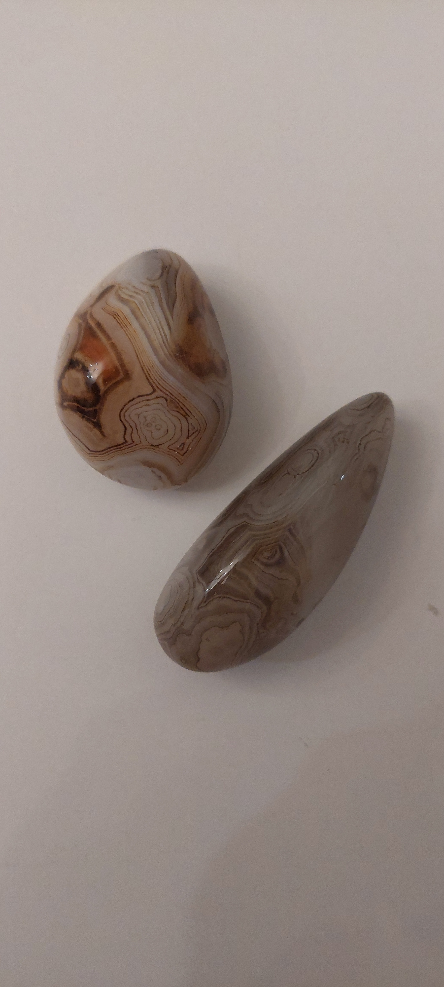 Two polished agate stones are placed on a plain, light-colored surface. One stone, on the left, is roughly teardrop-shaped and displays intricate banding in shades of brown, white, and orange. The other stone, to the right and slightly lower, is elongated and curved, with similar banding patterns in browns and grays. There is no indication of a specific location in Ivanovo, Russia, as the background is a nondescript surface without any identifying features.