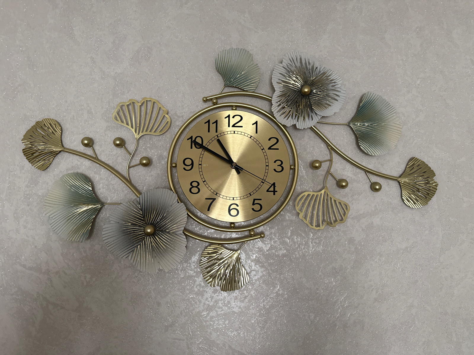 A circular, gold-colored analog clock is mounted on a textured, light gray wall. The clock face features black Arabic numerals from 1 to 12 and black hour and minute hands. The clock is part of a larger wall decoration consisting of metallic gold and muted green leaf-shaped ornaments and flower-like embellishments arranged symmetrically around the clock. The overall aesthetic suggests an art deco or botanical-inspired design. The image is a close-up, centered view of the clock and its surrounding decorative elements. No people, animals, or specific city locations are visible.