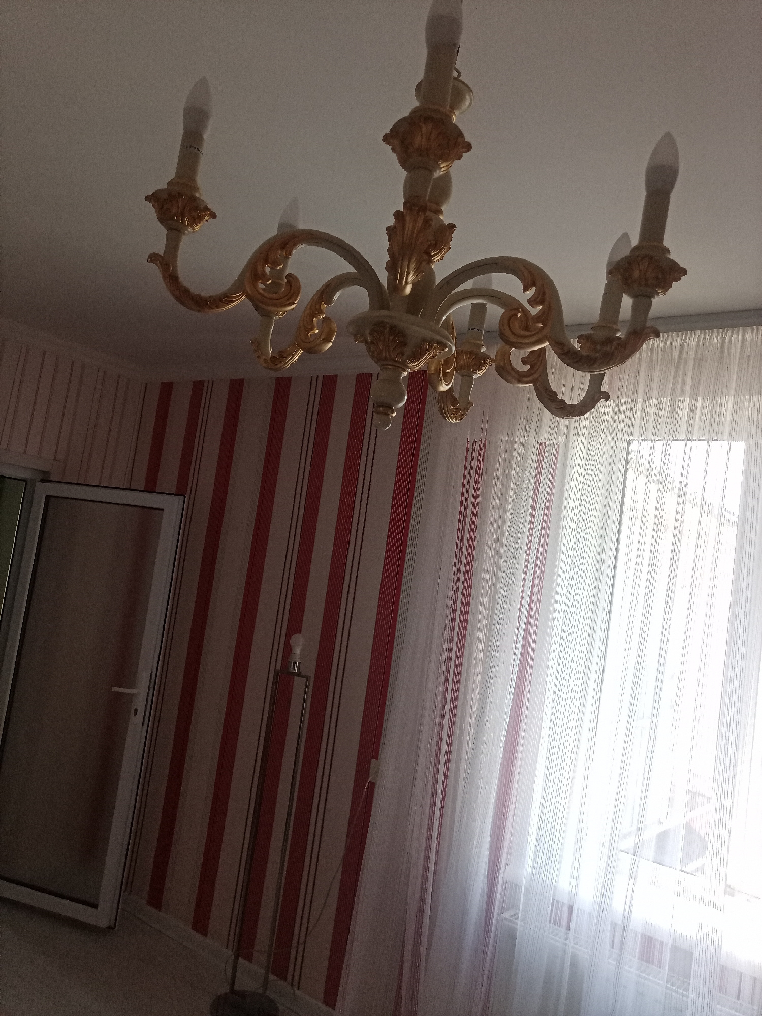 The image depicts an interior scene, likely a living room, with a chandelier as the focal point. The chandelier is ornate, with a cream-colored base and gold detailing, and has multiple candle-shaped light bulbs. The walls are adorned with striped wallpaper in shades of pink and red. To the left, a door with a glass panel is visible, and to the right, sheer white curtains hang over a window, allowing a bright, diffused light to enter the room. A tall floor lamp with a single bulb stands near the wall, to the left of the curtains. The floor appears to be carpeted or tiled. The overall impression is of a well-lit, furnished room. The location is identified as Durlești, Moldova. There are no people present in the image.