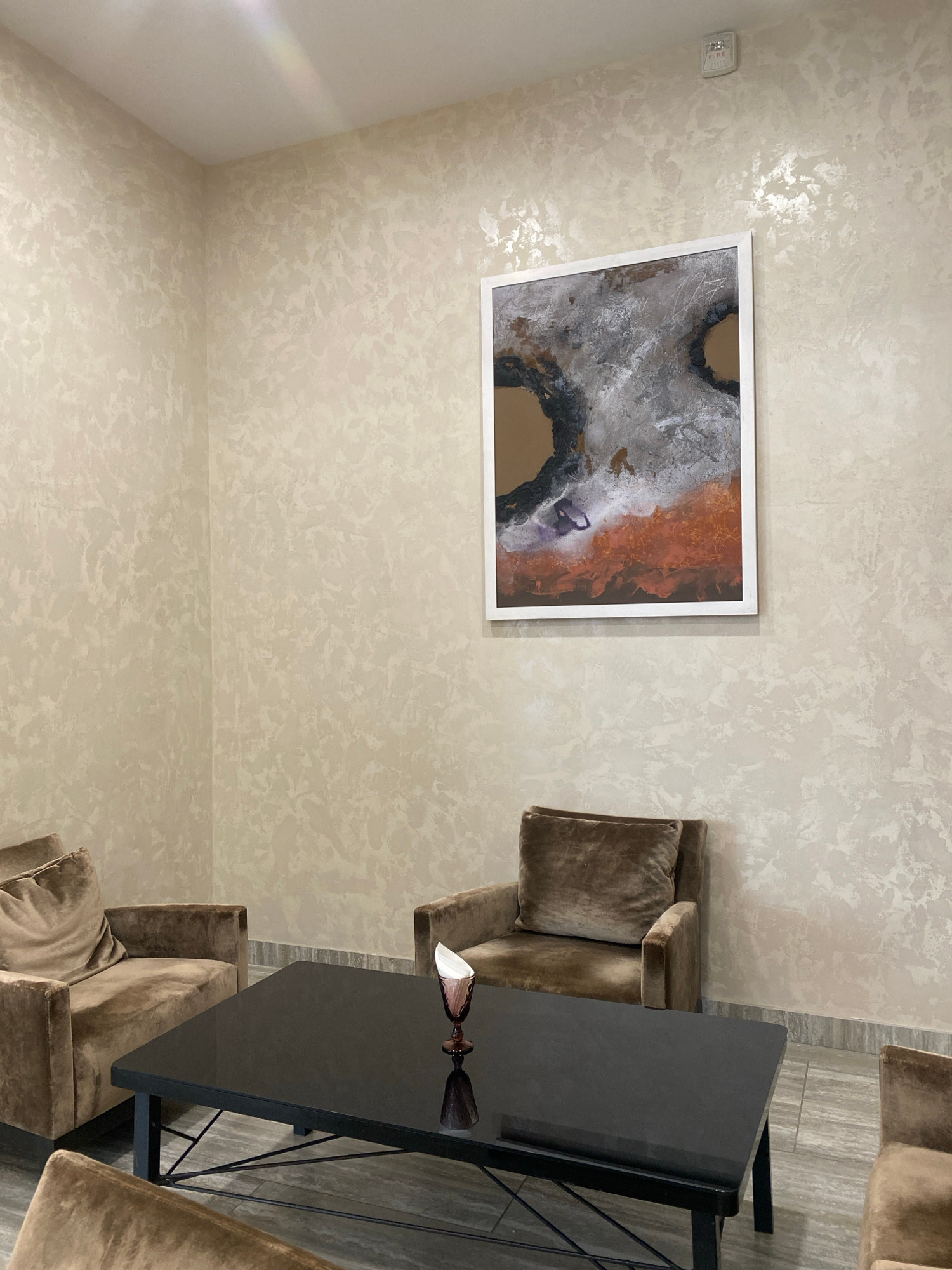 In an indoor setting, possibly a lounge or waiting area, a dark, rectangular coffee table with a black glossy surface is centrally positioned. It rests on a black metal frame. On the table, a single decorative glass with a narrow stem holds a white folded napkin. The table is flanked by two plush, taupe-colored armchairs, each with a square cushion. A third armchair is partially visible in the foreground, and another chair is visible to the right, also in taupe. The walls are textured with a pale beige finish, and a framed abstract artwork hangs on the wall above the armchairs. The artwork features a white frame and a composition of earthy tones, including browns, grays, and oranges, with darker circular elements. A white fire alarm is mounted on the upper right portion of the wall. The flooring appears to be made of large, dark gray tiles.