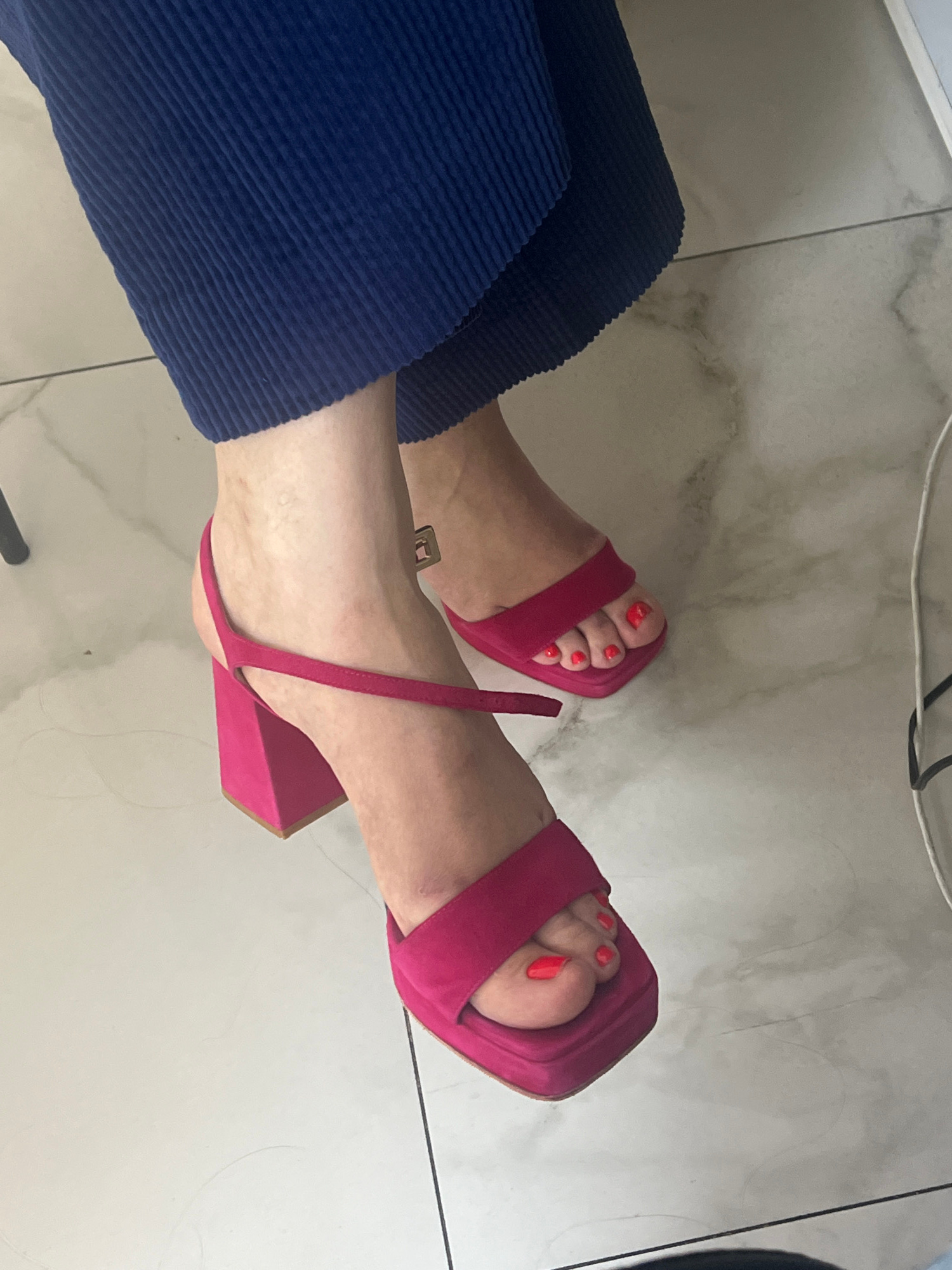 A person is wearing bright pink platform sandals and navy blue corduroy pants. The sandals have a square toe and a thin strap that goes around the ankle. The person's toenails are painted a vibrant coral color. They are sitting on a floor with a marble-like pattern, possibly indoors. The lighting suggests it is daytime. There are no visible text elements in the image. The scene is a close-up shot focused on the person's lower legs and feet.