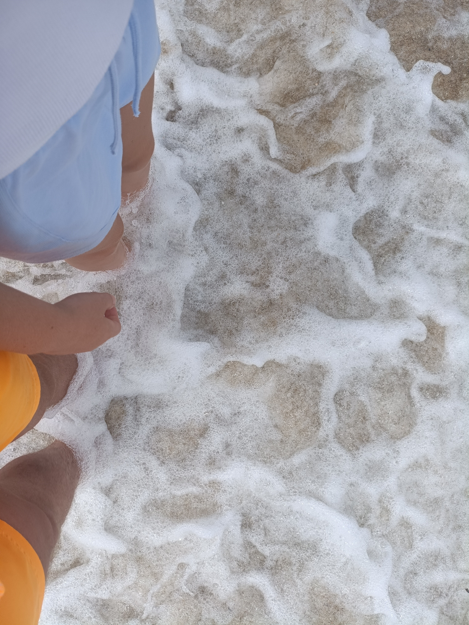 The image shows the lower bodies of two people standing at the edge of the sea, with waves lapping around their feet. One person is wearing light blue shorts and a white top, while the other is wearing bright orange shorts. The sand beneath the water is visible through the white foam of the waves. The scene appears to be a casual day at the beach, possibly during daylight hours, given the brightness of the scene. The image evokes a sense of relaxation and enjoyment of a beach environment. There is no visible text in the image. The location is identified as Sagunto, Spain.