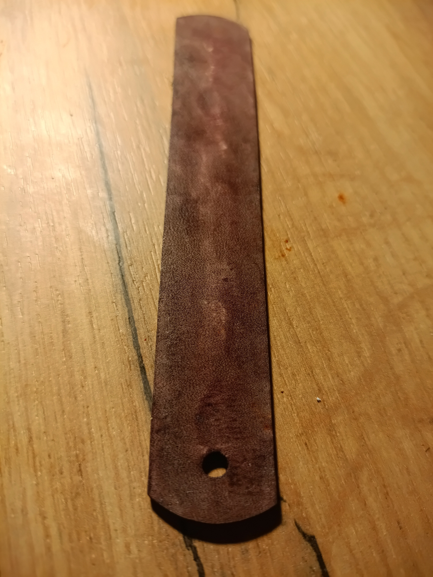 A single, dark brown, rectangular piece of leather with rounded corners lies on a light brown wooden surface. There is a small, circular hole near the bottom edge of the leather. The wooden surface exhibits a prominent grain pattern and appears to be a table or workbench. There are no people or other objects visible in the image. The location in Irkutsk, Russia is not discernible from the image.