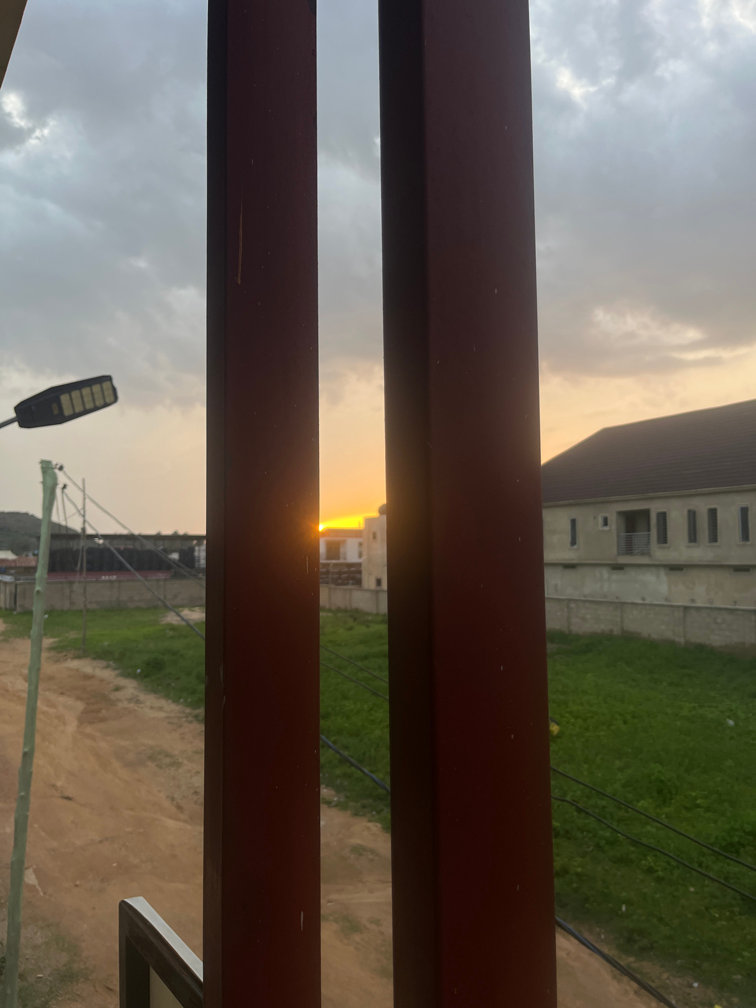 An outdoor, eye-level, medium shot shows a hazy sunset viewed between two dark red, rectangular vertical posts. The sky is overcast with white and grey clouds. In the distance, a bright orange sun peeks through a gap in the clouds, illuminating the sky with yellow and orange hues. To the left of the frame, a modern, black street lamp with LED lights is mounted on a thin pole, extending from the left edge of the image toward the center. Beyond the lamp, a grey concrete fence stretches across the middle ground, separating a dirt road from a field of green grass. Several electrical wires are strung across the field. In the background, on the right side, a light-colored, multi-story building with a dark brown roof stands behind the fence. The building has several rectangular windows and a balcony on the upper floor. The scene is set in Gura Top, Nigeria.