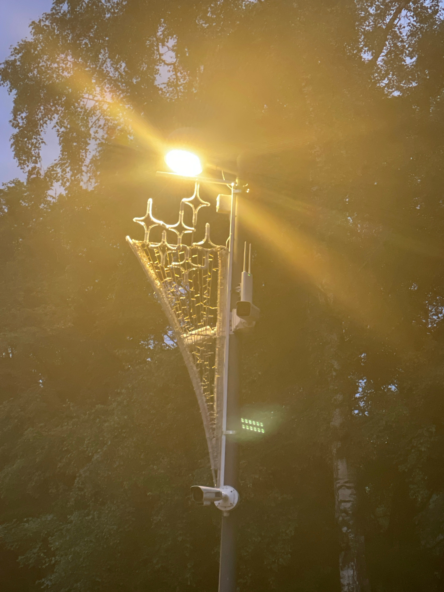 The image depicts a street lamp with decorative elements and a security camera mounted on a pole. The lamp is illuminated, casting a bright yellow light and creating a lens flare effect that dominates the upper portion of the image. The decorative elements consist of a series of star-shaped figures and what appears to be a mesh-like structure adorned with small lights. Below the decorative elements, a security camera is visible, along with another device that might be a communication antenna. The background is filled with the foliage of trees, suggesting an outdoor environment. The lighting indicates it is either dusk or nighttime, or the street lamp is switched on during the day. The overall scene appears to be in an urban setting, possibly a park or a street. There are no people visible in the image.